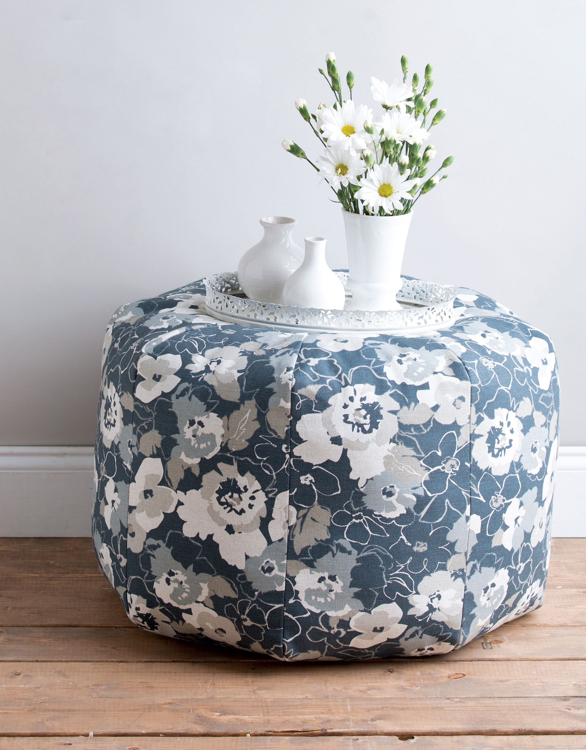The Sewing Tuffets & Poufs by Leisure Arts, featuring blue and white floral fabric inspired by classic designs, sits on a wooden floor with a white vase of daisies, two small vases, and a tray against a light gray wall.