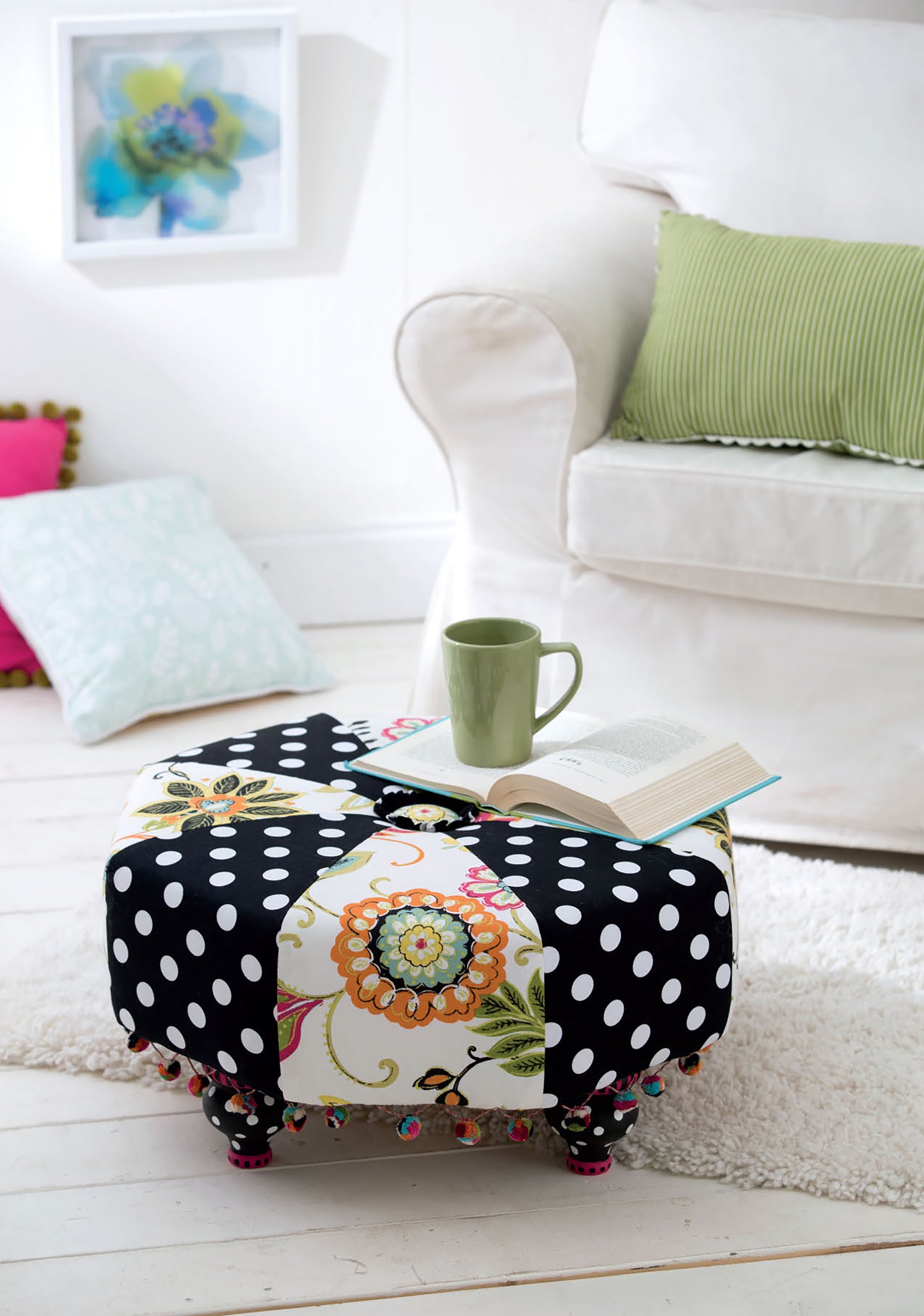 The Sewing Tuffets & Poufs by Leisure Arts, with black and white polka dots and floral patterns, sits in a bright living room holding an open book and green mug. Nearby are a white armchair with a green pillow and two floor cushions.