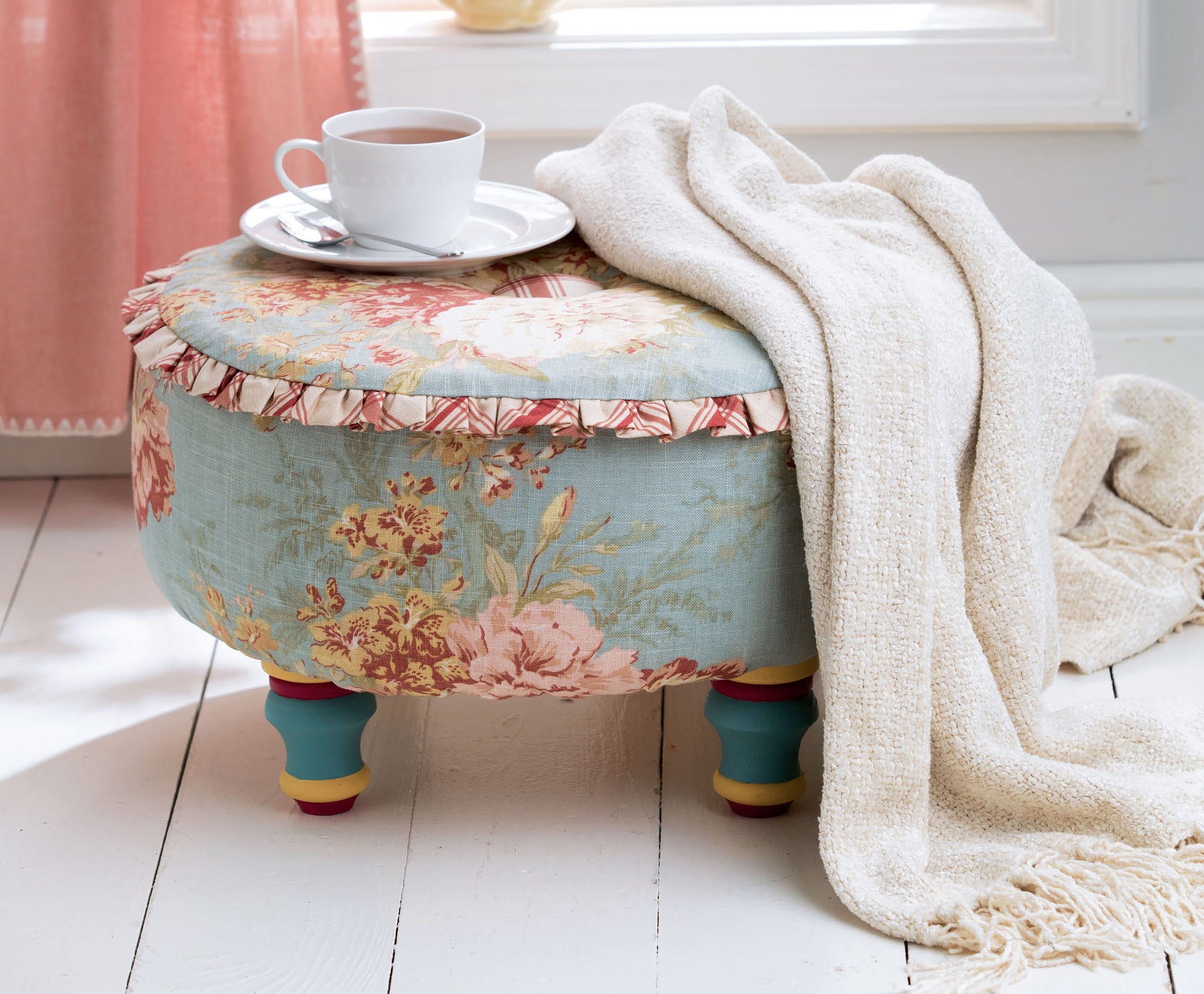 The Sewing Tuffets & Poufs by Leisure Arts features a floral upholstered ottoman with colorful legs, detailed pouf patterns, topped with a white teacup and saucer, and draped in a cream knit blanket on a white floor near a pink curtain.