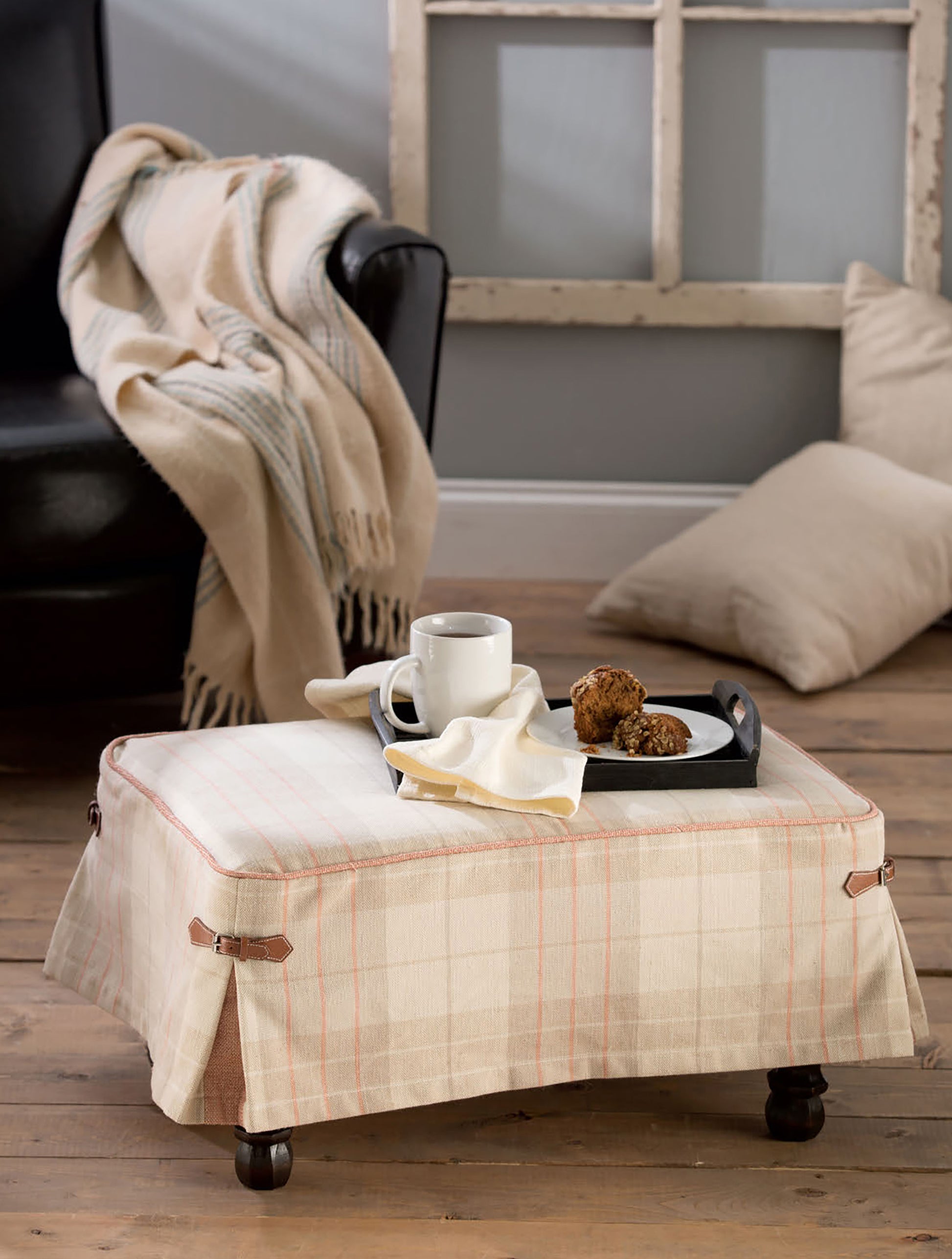 A cozy living room features a Leisure Arts Sewing Tuffets & Poufs plaid ottoman holding a tray with a white mug, napkin, and snacks; in the background, a black chair with striped throw and beige pillow sits on the wooden floor.