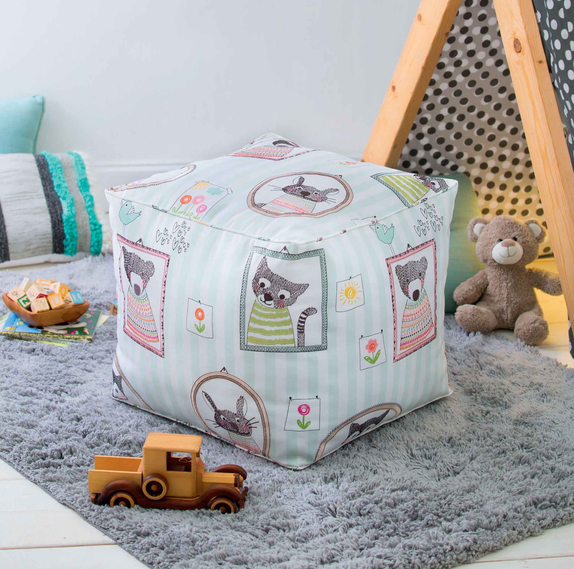 A cozy children's playroom with a soft gray rug, Leisure Arts Sewing Tuffets & Poufs featuring animal prints and playful patterns, a teepee tent, teddy bear, cushions, wooden toy truck, and building blocks.