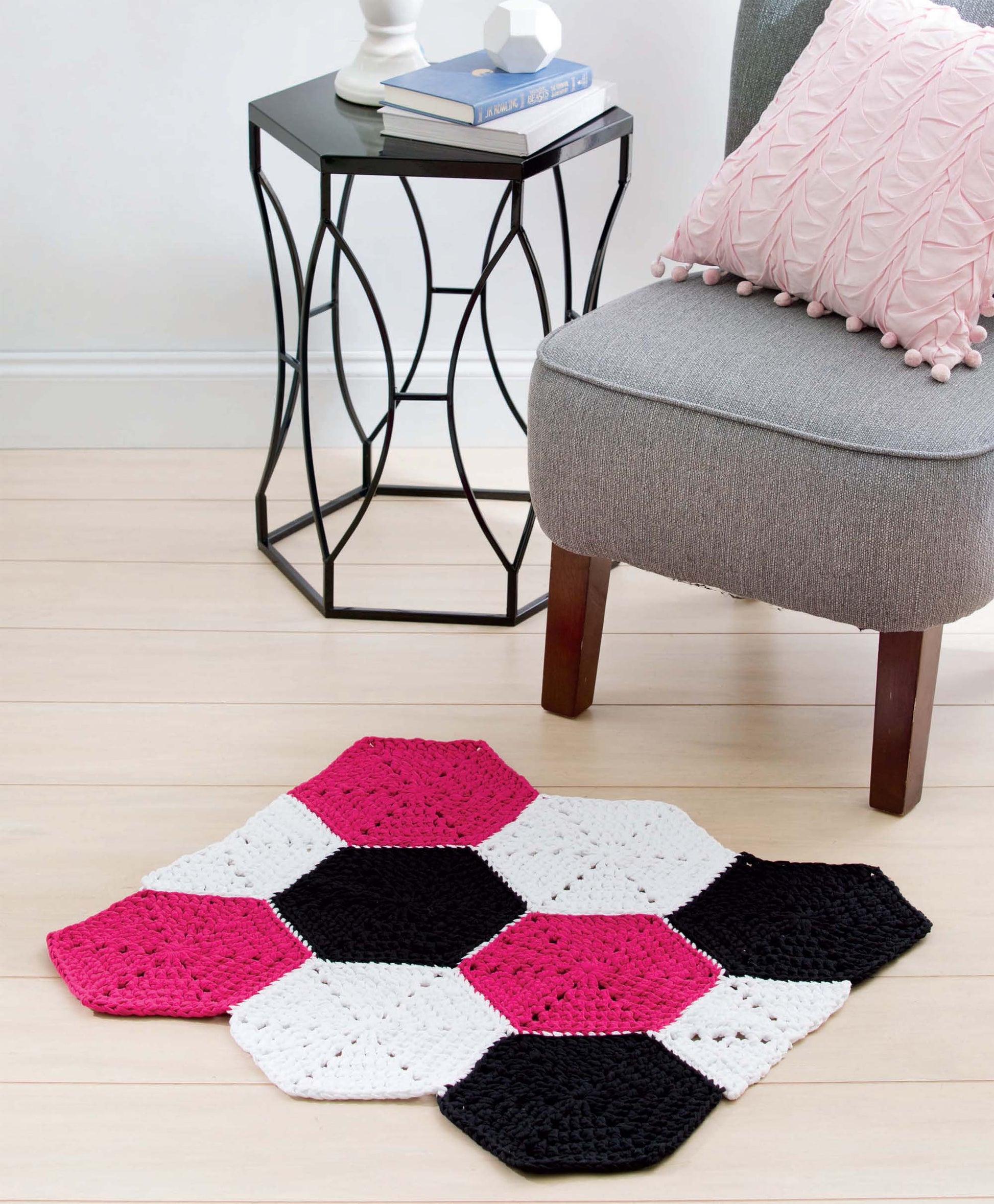 A modern T-Shirt Yarn Home Décor Crochet rug from Leisure Arts, featuring pink, black, and white hexagons, is placed on a light wood floor beside a gray chair with a pink pillow and a sleek black metal side table with books.