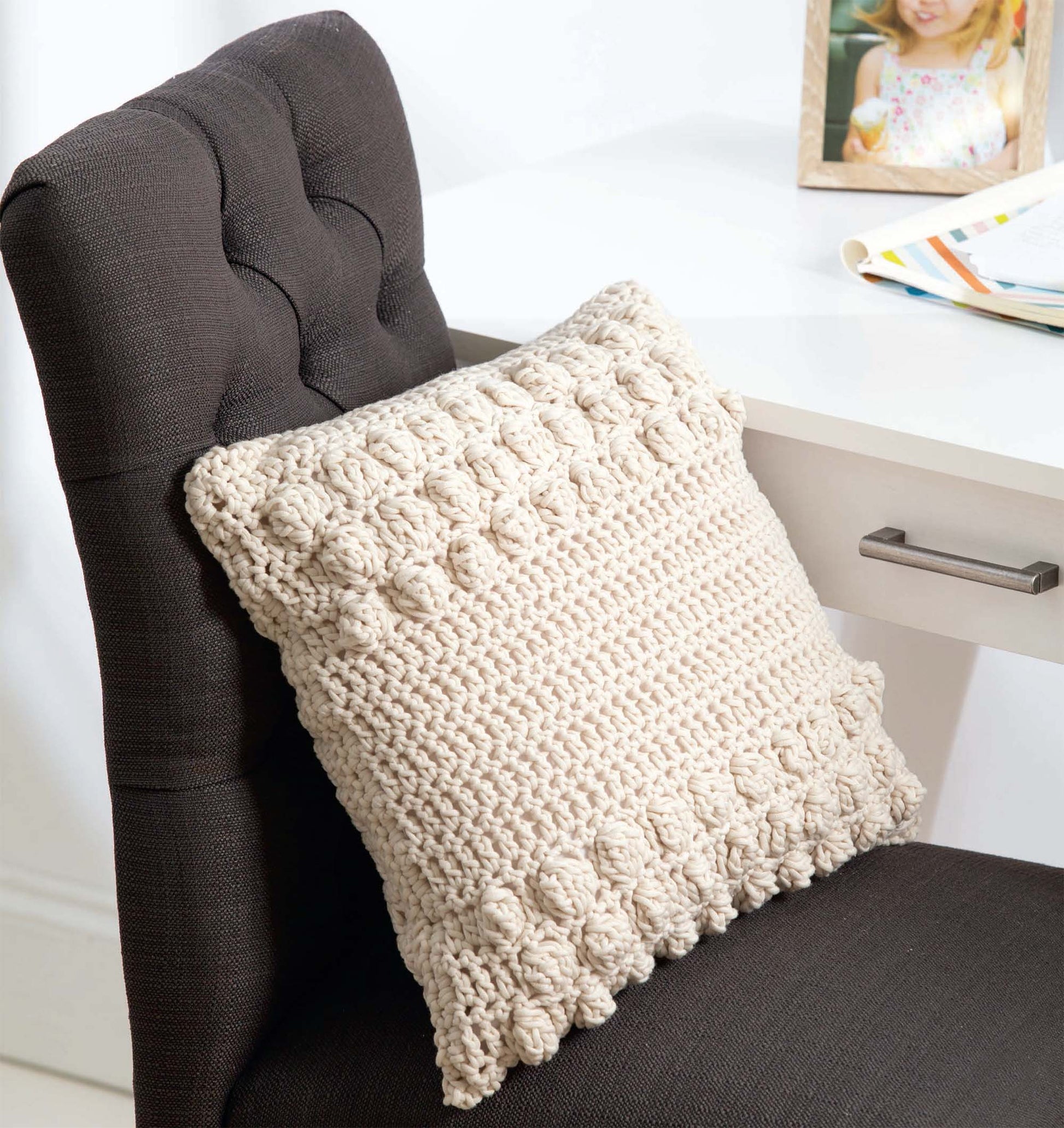 A cozy crochet pillow made using the T-Shirt Yarn Home Décor Crochet Digital Download by Leisure Arts rests on a dark chair beside a white desk, with an open book, framed photo, and a basket set nearby for a stylish touch.