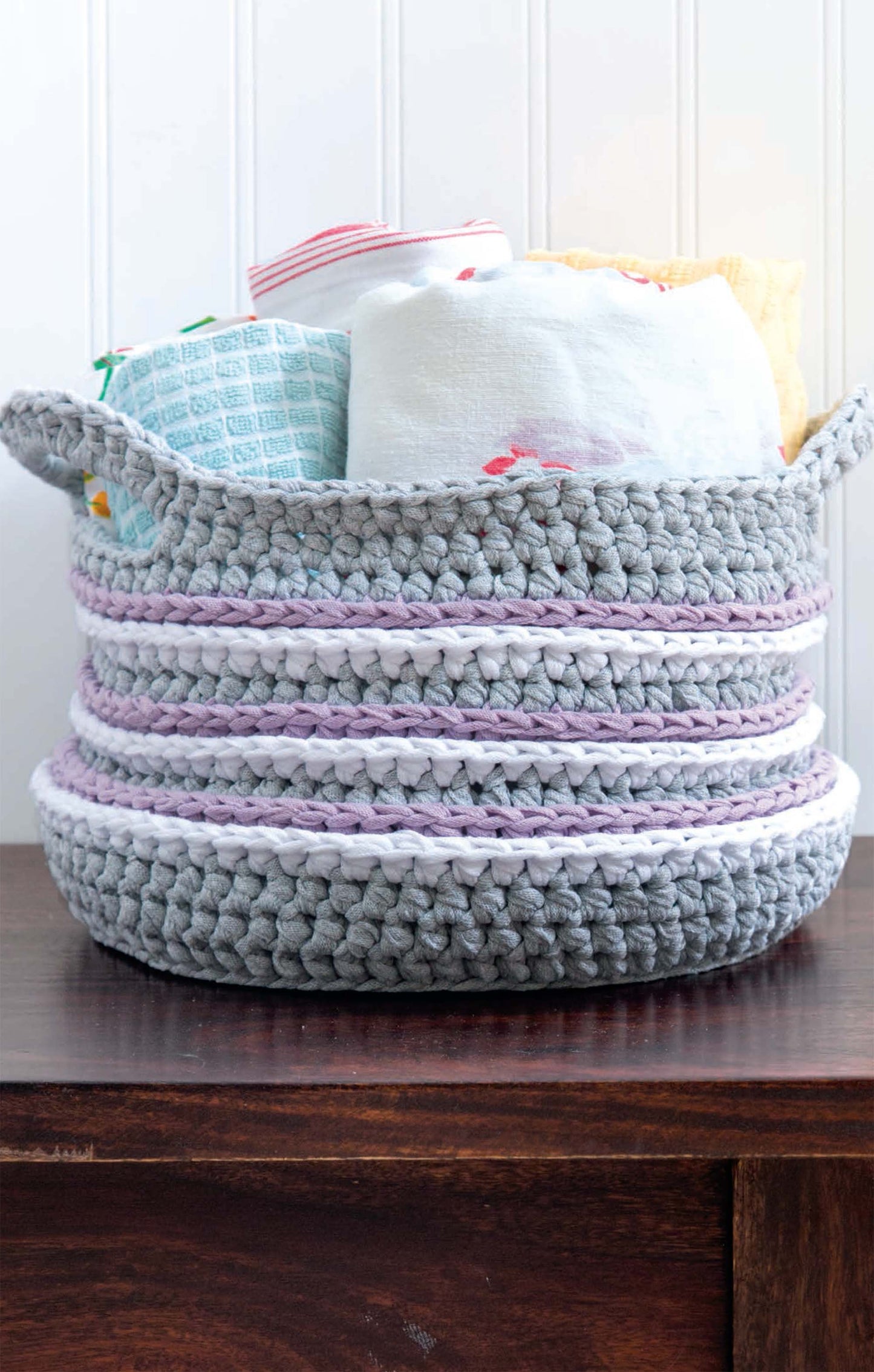 A gray basket with white and lavender stripes, made using Leisure Arts' T-Shirt Yarn Home Décor Crochet Digital Download, holds folded towels and rests on a dark wooden surface against a white paneled wall.