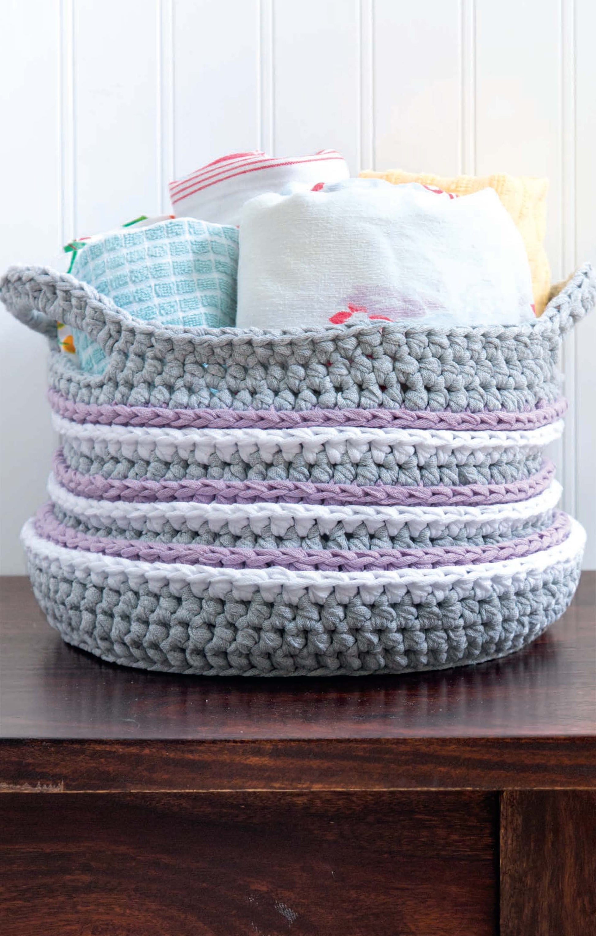A gray basket with white and lavender stripes, made using Leisure Arts' T-Shirt Yarn Home Décor Crochet Digital Download, holds folded towels and rests on a dark wooden surface against a white paneled wall.