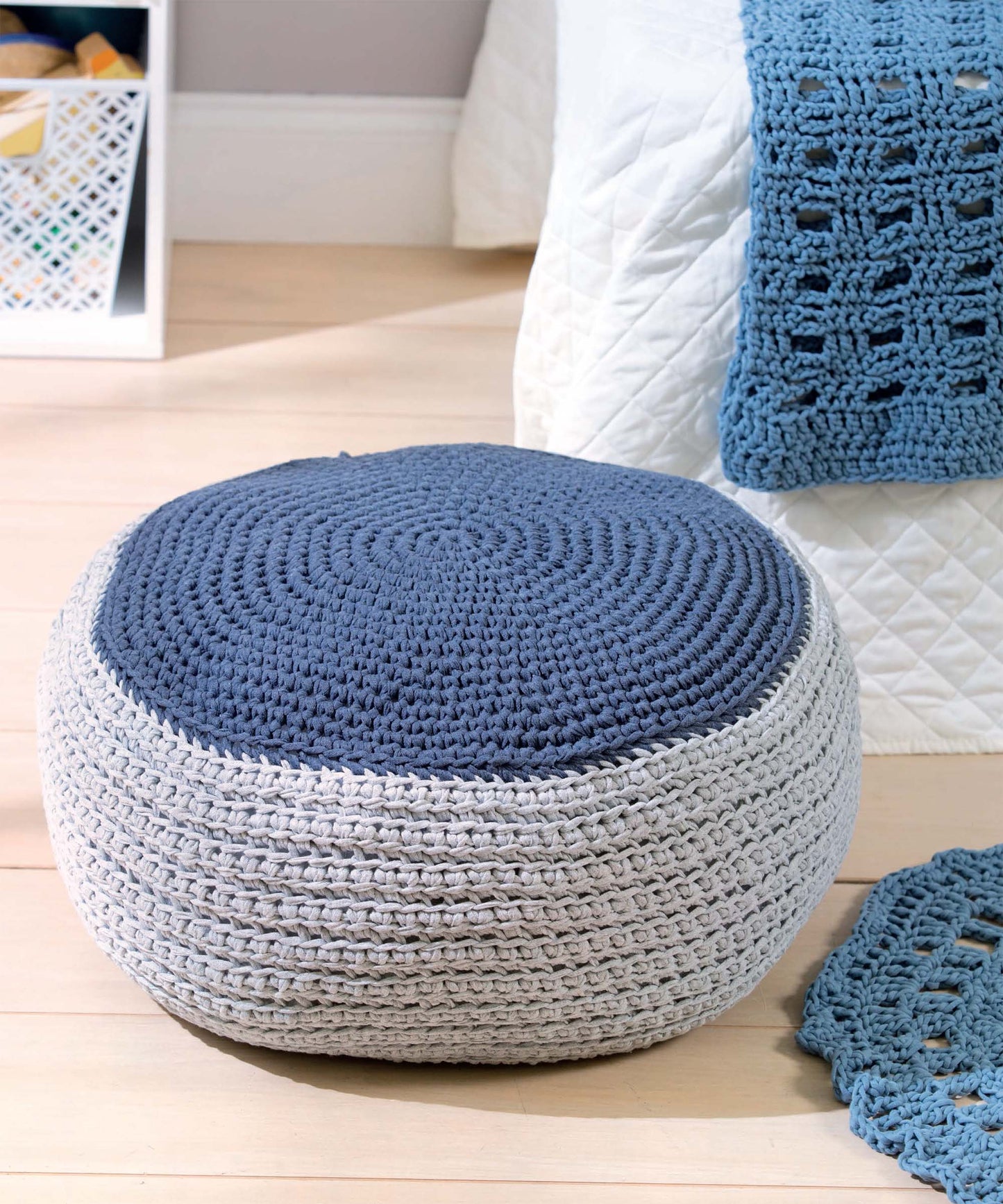 A round crochet pouf, made with the T-Shirt Yarn Home Décor Crochet Digital Download by Leisure Arts, features a blue top and light gray sides. It sits on a wooden floor next to a bed with a white quilt and openwork throw.