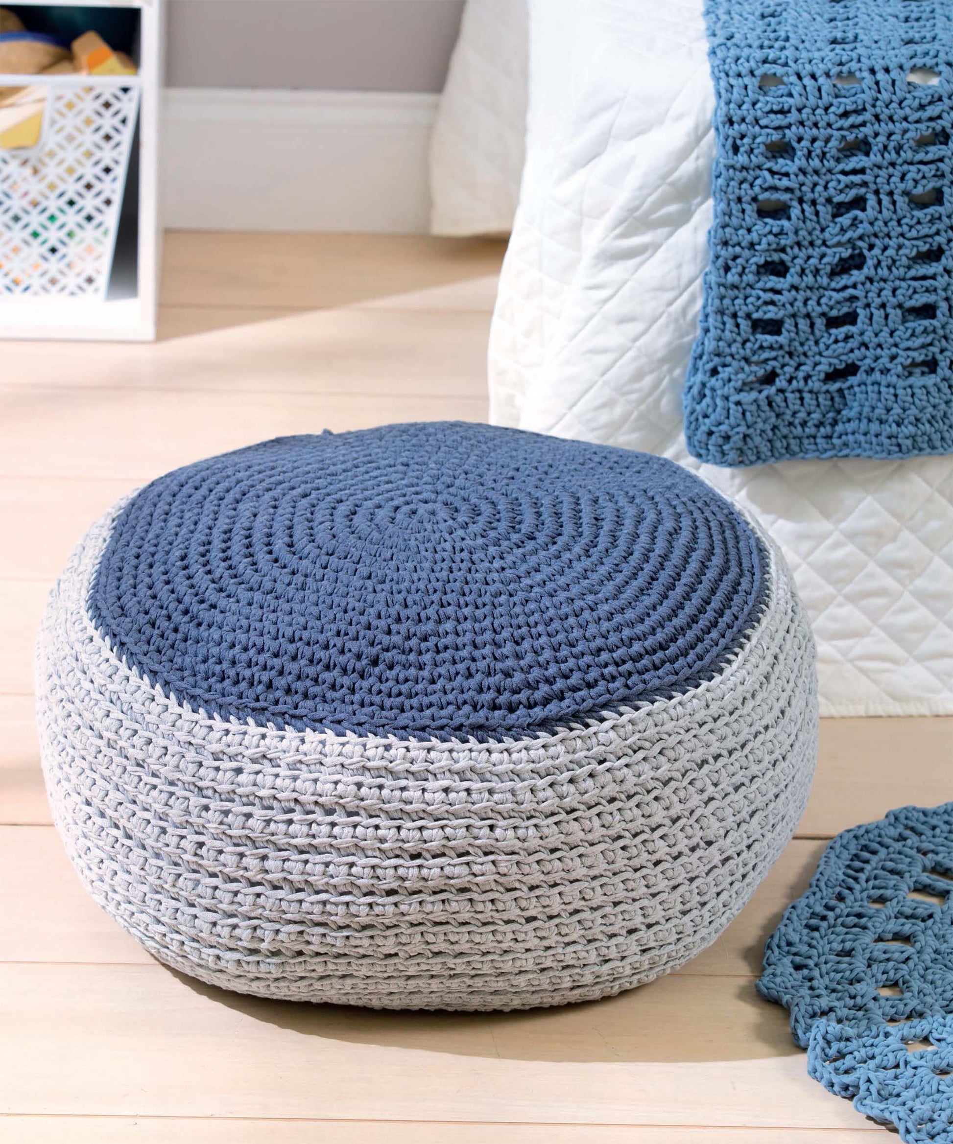 A round crochet pouf, made with the T-Shirt Yarn Home Décor Crochet Digital Download by Leisure Arts, features a blue top and light gray sides. It sits on a wooden floor next to a bed with a white quilt and openwork throw.