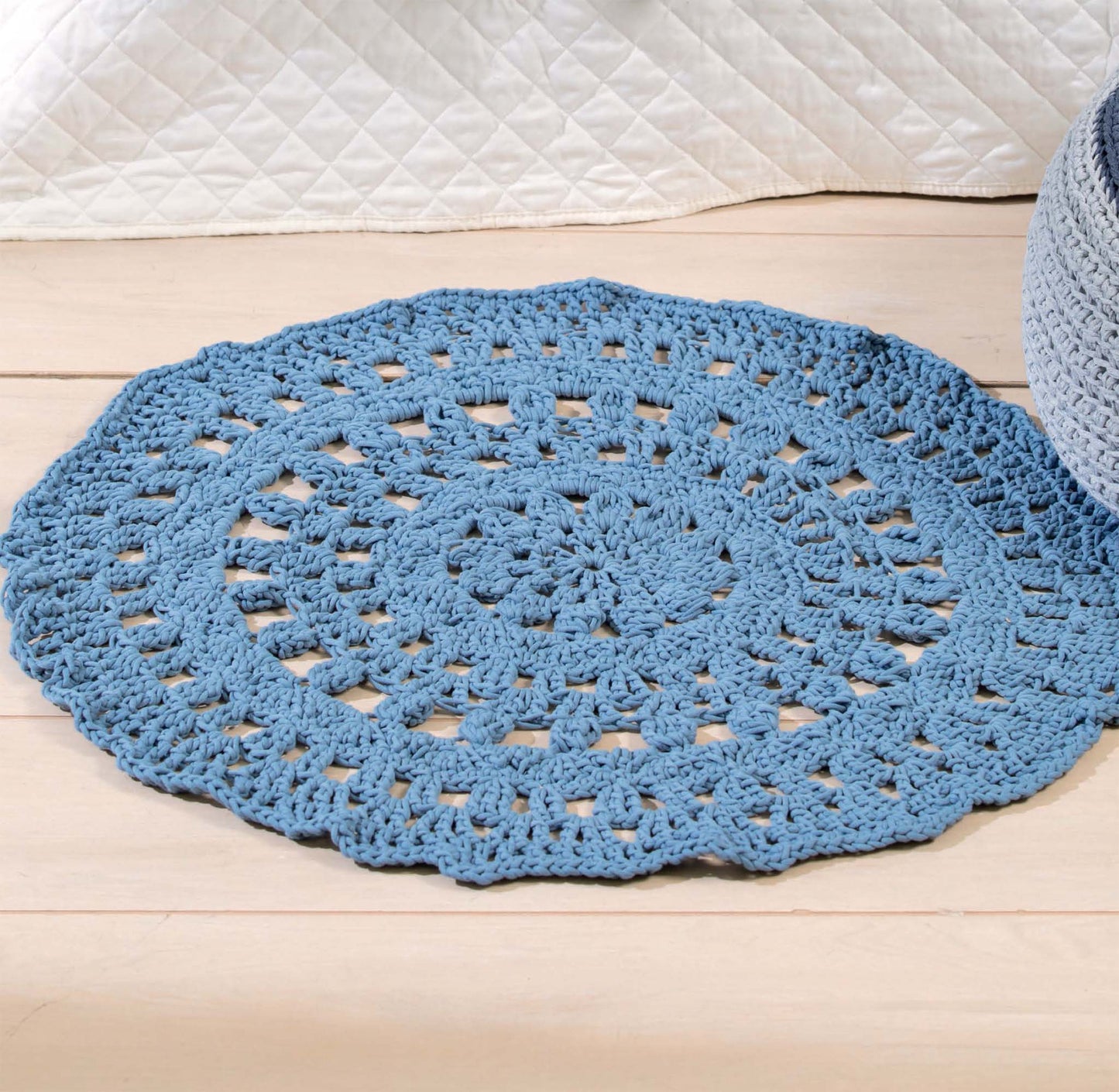 A round, blue lacy-patterned rug from Leisure Arts’ T-Shirt Yarn Home Décor Crochet Digital Download is displayed on a light wood floor beside a white quilted blanket and an openwork throw.