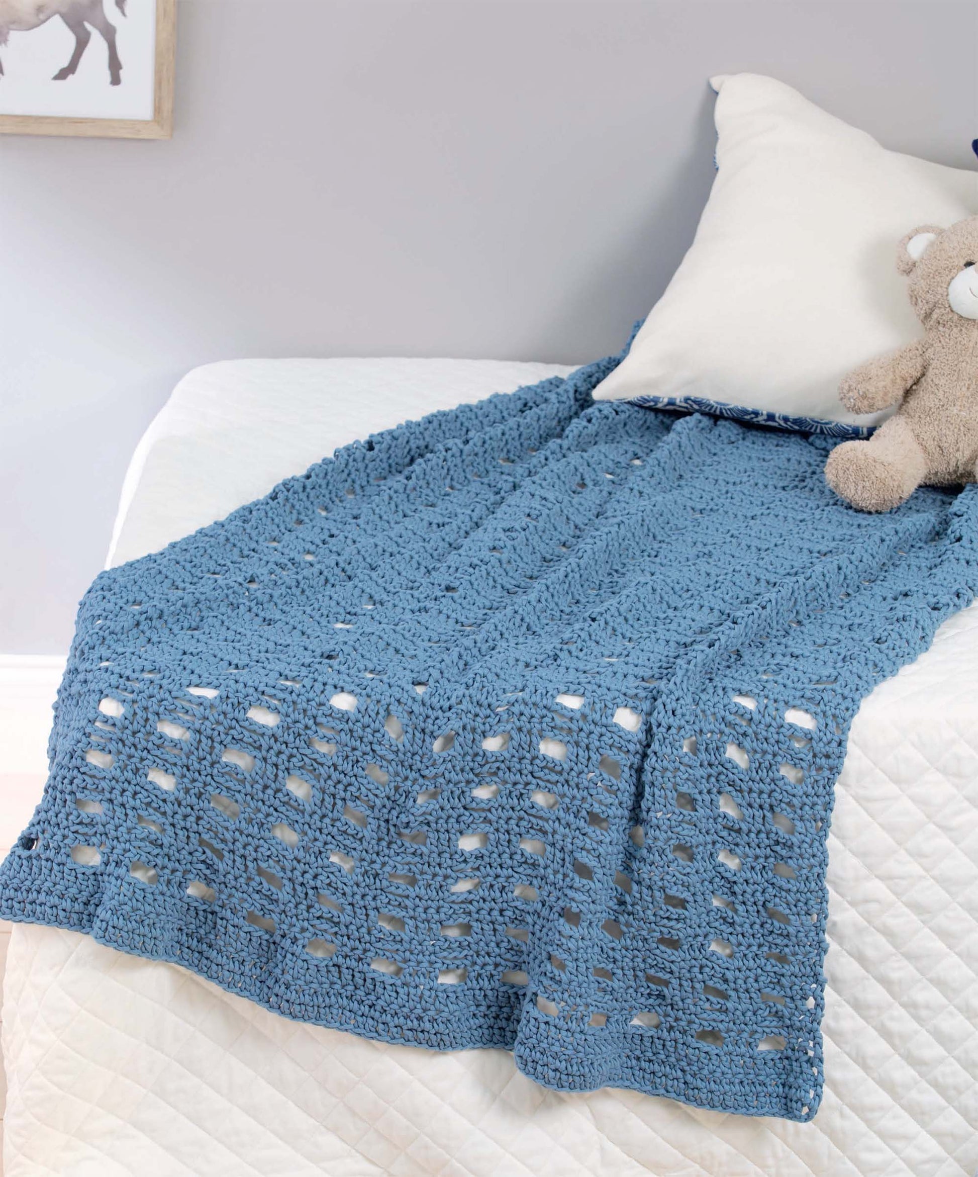 A crochet throw made with the T-Shirt Yarn Home Décor Crochet Digital Download by Leisure Arts is draped over a bed with a white pillow and stuffed bear, next to a hexagon motif rug, against a light gray wall.
