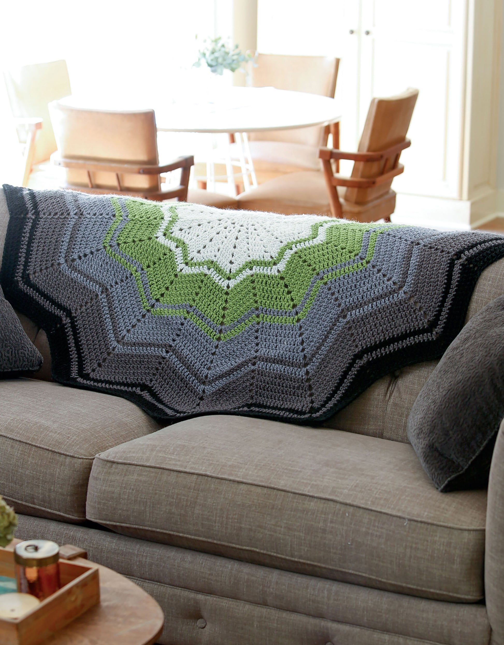 The Stripe It Your Way Crochet for Home & Fashion blanket by Leisure Arts, in black, gray, green, and white chevrons, is draped over a beige sofa with a gray pillow and wooden coffee table in a bright living room.