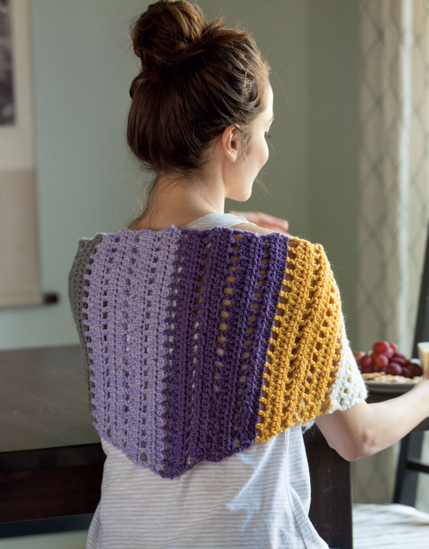 A woman with brown hair in a bun models a striped, lacy crochet wrap in gray, purple, and yellow from Leisure Arts' "Stripe It Your Way Crochet for Home & Fashion," offering perfect beginner inspiration as she sits at a sunlit table with grapes.