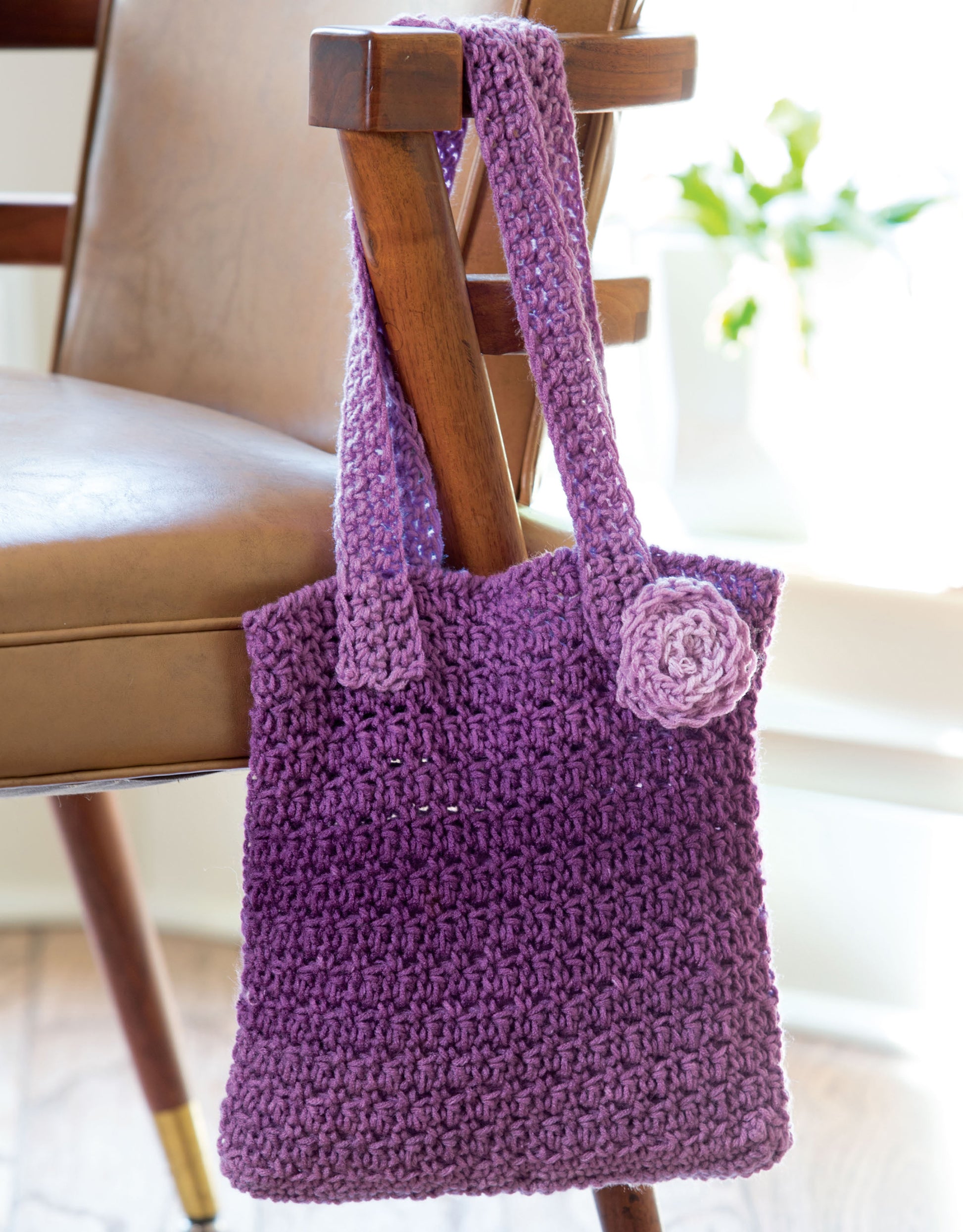 The Stripe It Your Way Crochet for Home & Fashion bag by Leisure Arts, featuring a purple crochet design and flower detail, hangs on a wooden chair with a tan leather seat. A blurred green plant appears in the background.