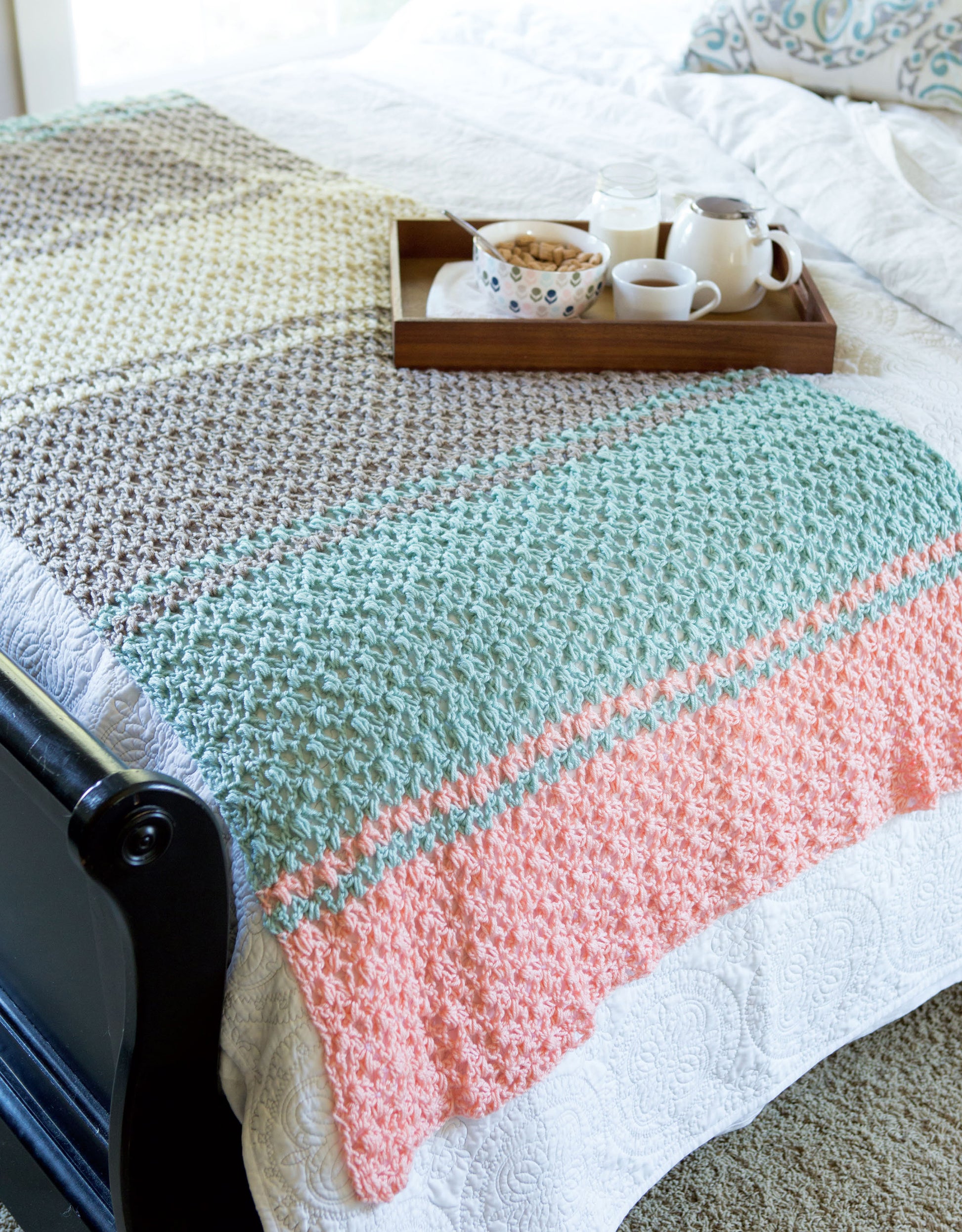 A tray with tea, milk, and cereal rests on a bed topped with white bedding and a dark wood footboard, featuring a peach, mint, gray, and cream star blanket inspired by Stripe It Your Way Crochet for Home & Fashion by Leisure Arts.