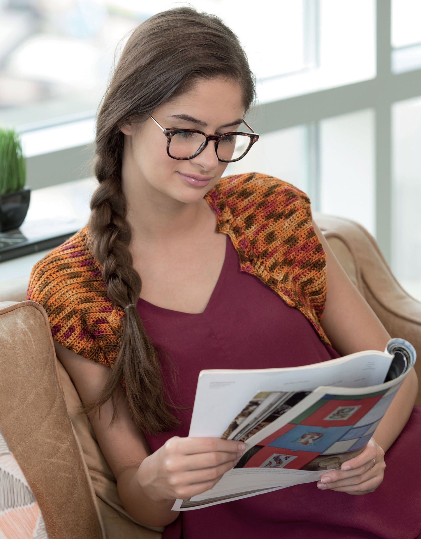 A woman with long brown hair in a braid, wearing glasses, a burgundy dress, and a Leisure Arts Crochet Shrugs & Boleros piece, sits on a sofa reading a magazine in a bright room.