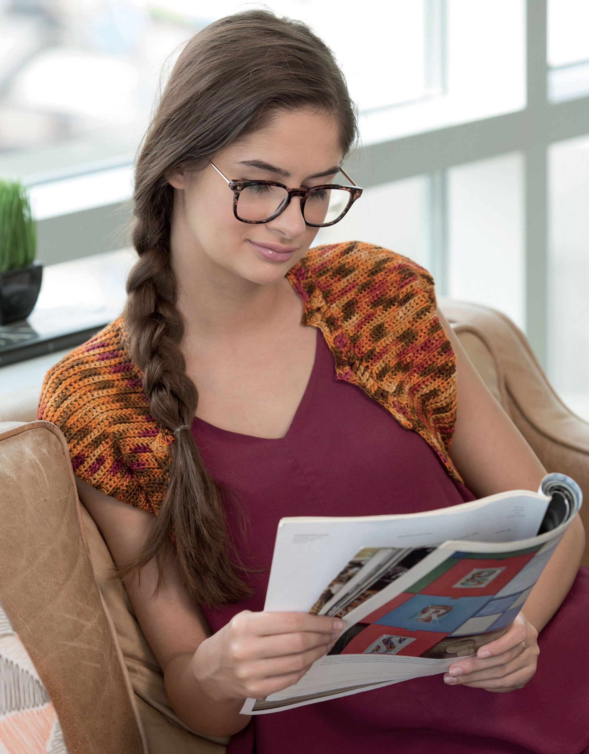A woman with long brown hair in a braid, wearing glasses, a burgundy dress, and a Leisure Arts Crochet Shrugs & Boleros piece, sits on a sofa reading a magazine in a bright room.