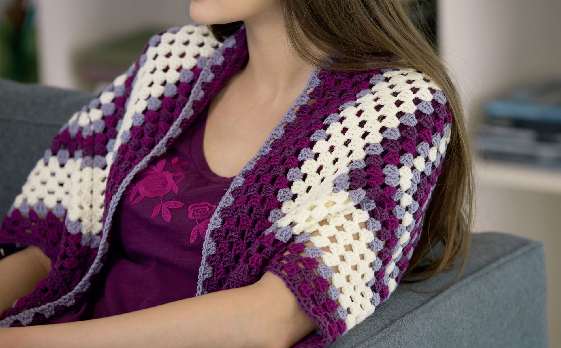 A woman with long brown hair sits on a gray couch in a purple floral shirt and a striped shawl inspired by Crochet Shrugs & Boleros from Leisure Arts. Her face is partially out of frame.
