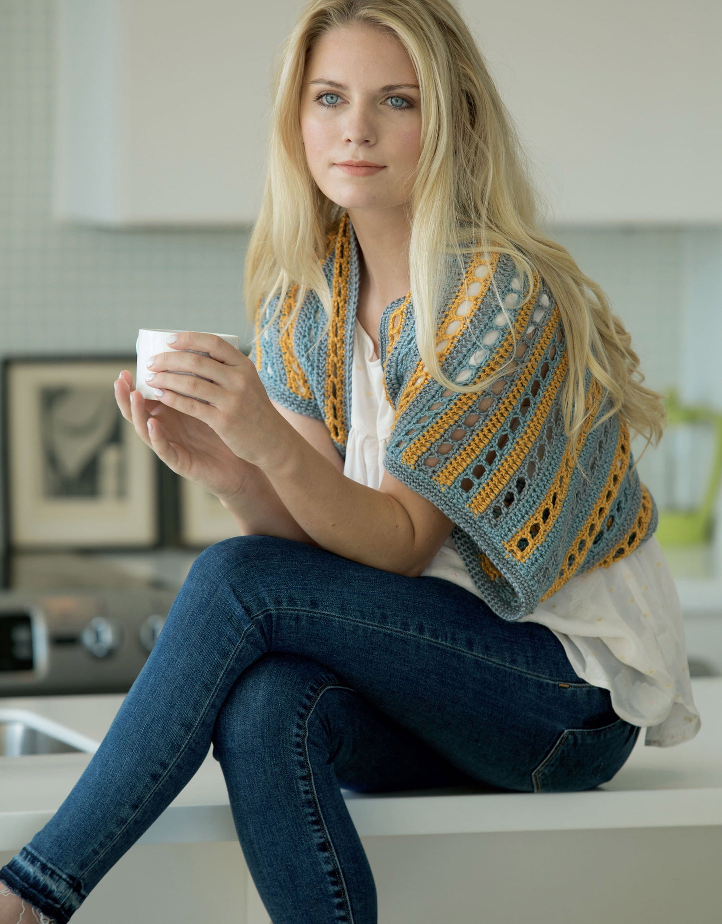 A woman with long blonde hair sits on a kitchen counter, holding a white mug. She wears blue jeans and a vibrant Crochet Shrugs & Boleros by Leisure Arts, featuring grey, blue, and yellow stripes—ideal for beginners.