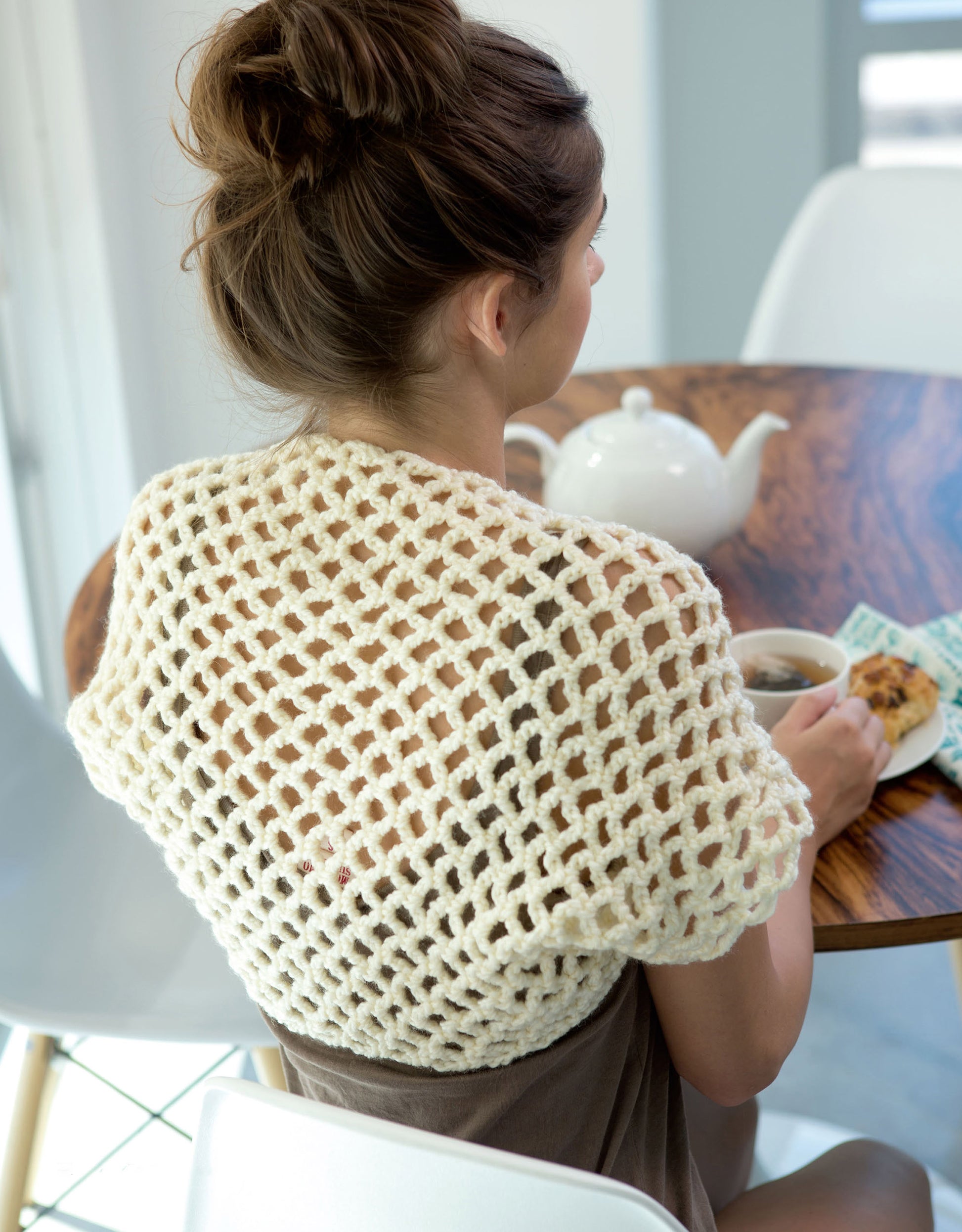A woman with brown hair in a bun sits at a wooden table, wearing a cream open-knit shrug from Leisure Arts’ Crochet Shrugs & Boleros. She holds a cup of tea beside a white teapot, plate of cookies, and blue napkin.