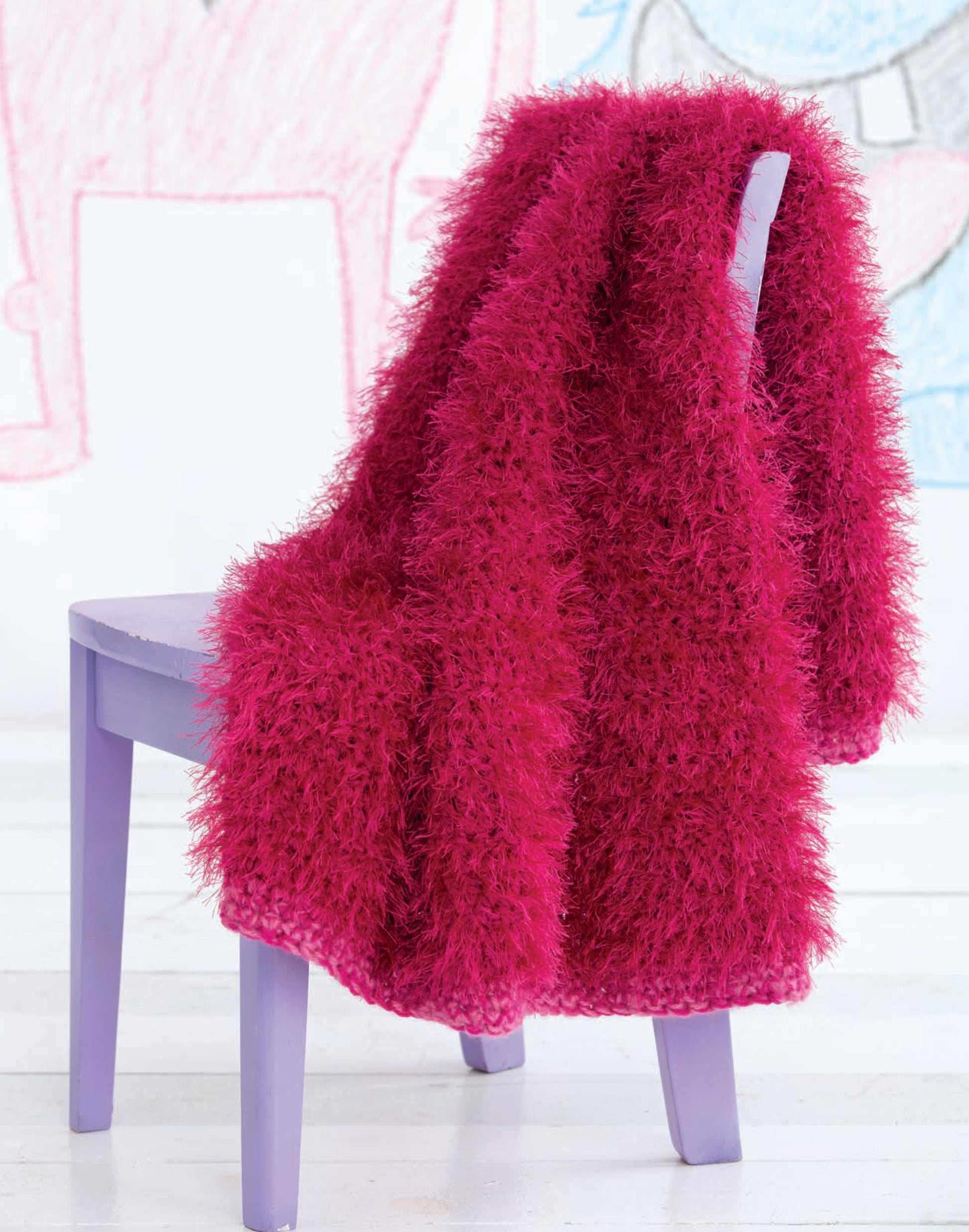 A bright pink, easy-to-crochet Leisure Arts Monster Playsets blanket drapes over a lavender chair, with animal drawings in the background enhancing the creative play vibe against a white wall.