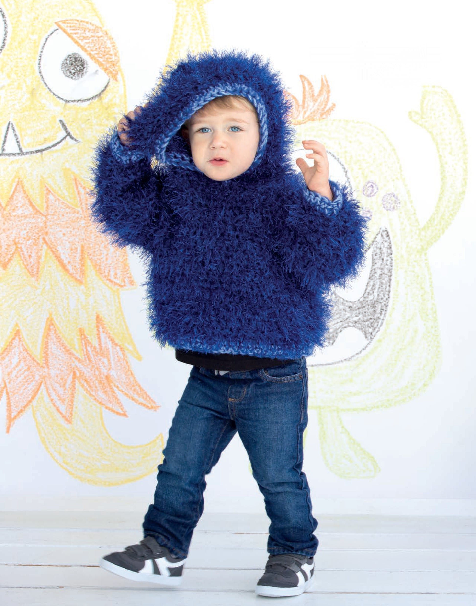 A young child in a fuzzy blue hoodie poses indoors by a wall of colorful monster drawings, reflecting the playful spirit of Leisure Arts’ Monster Playsets.