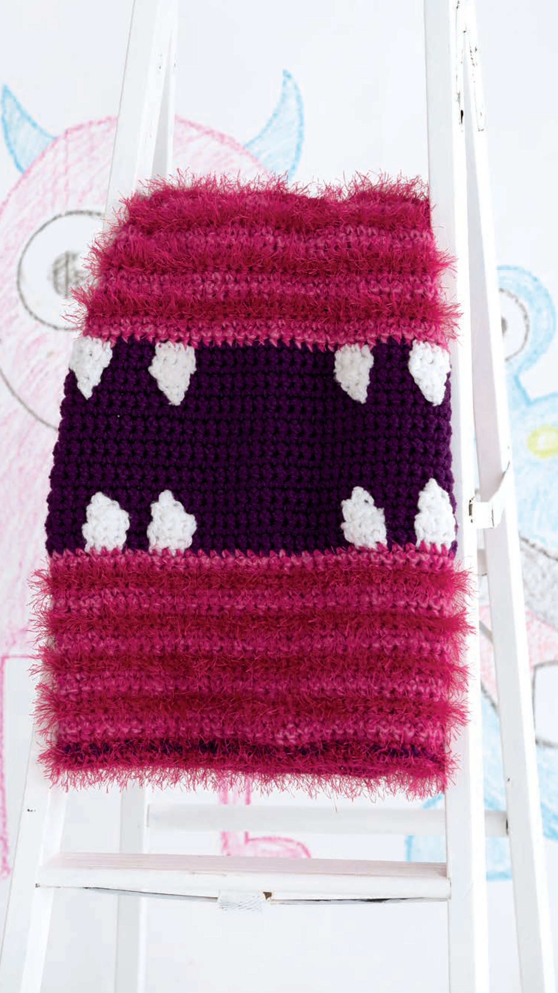 The Leisure Arts Monster Playsets features a knitted piece with fuzzy pink stripes and a central purple band with white, pointed monster teeth shapes, displayed on a white ladder—perfect for creative play blankets. Cartoon monster art appears in the background.