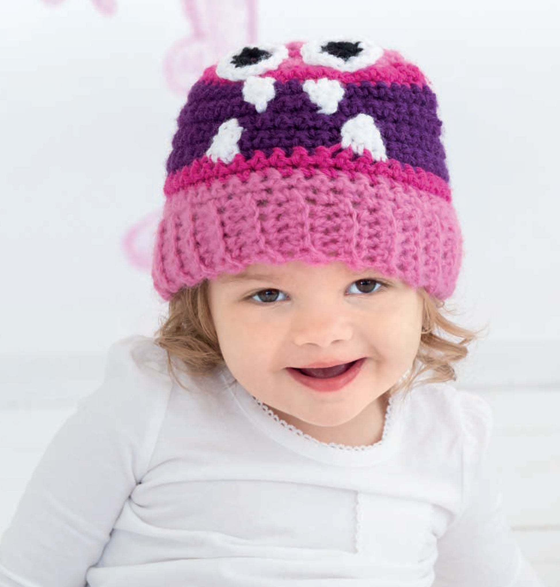 A smiling toddler models a pink and purple “Monster Playsets” hat by Leisure Arts, featuring big eyes and white teeth, while wearing a white shirt against a light background.