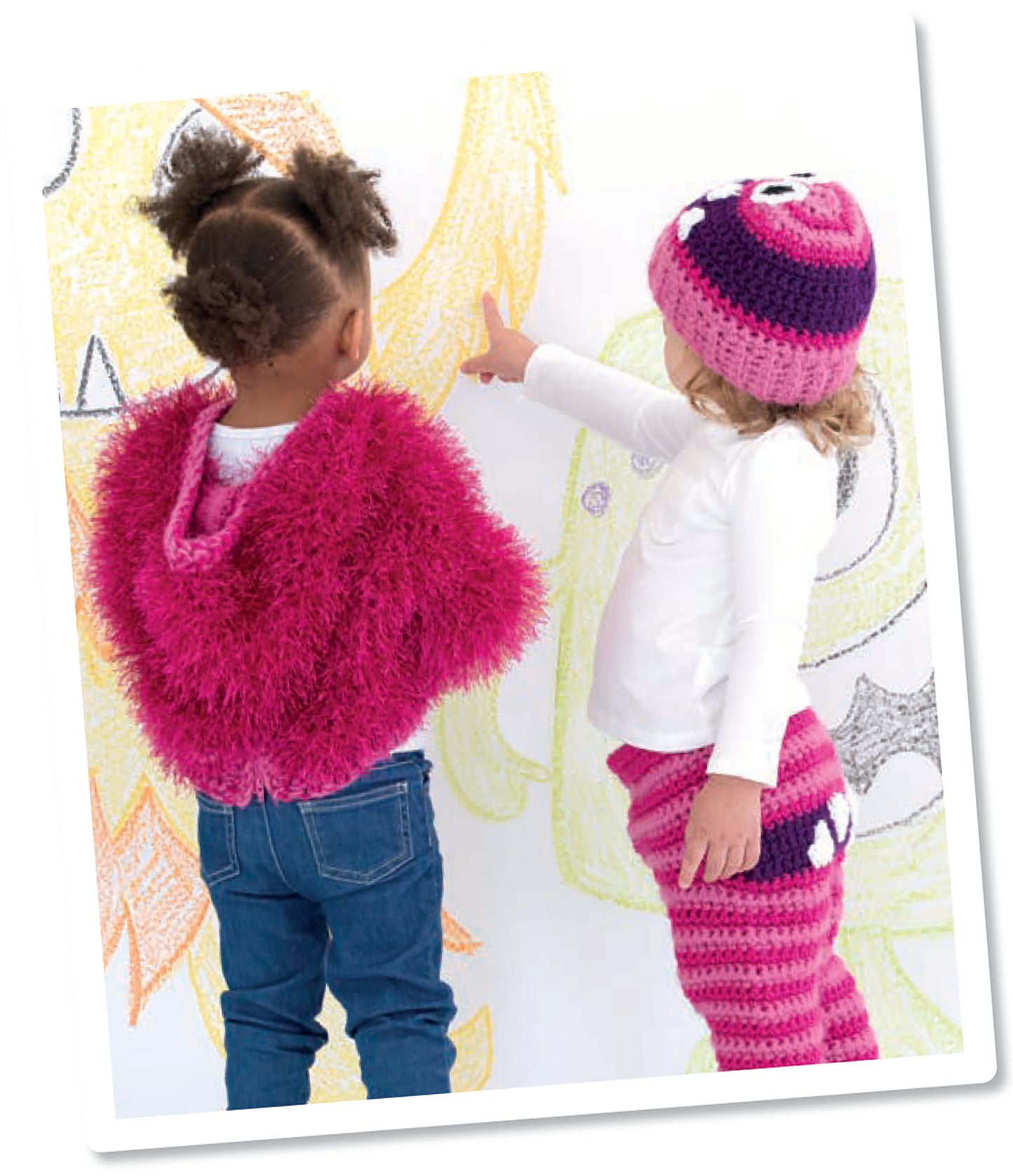 Two young children, dressed in easy-to-crochet outfits, stand by a wall of colorful crayon art. One points at the drawings. They’re enjoying playtime with Monster Playsets from Leisure Arts, adding creativity to their imaginative fun.