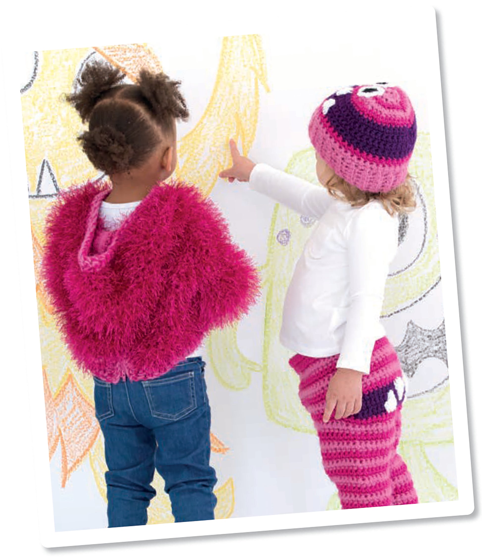 Two young children, dressed in easy-to-crochet outfits, stand by a wall of colorful crayon art. One points at the drawings. They’re enjoying playtime with Monster Playsets from Leisure Arts, adding creativity to their imaginative fun.