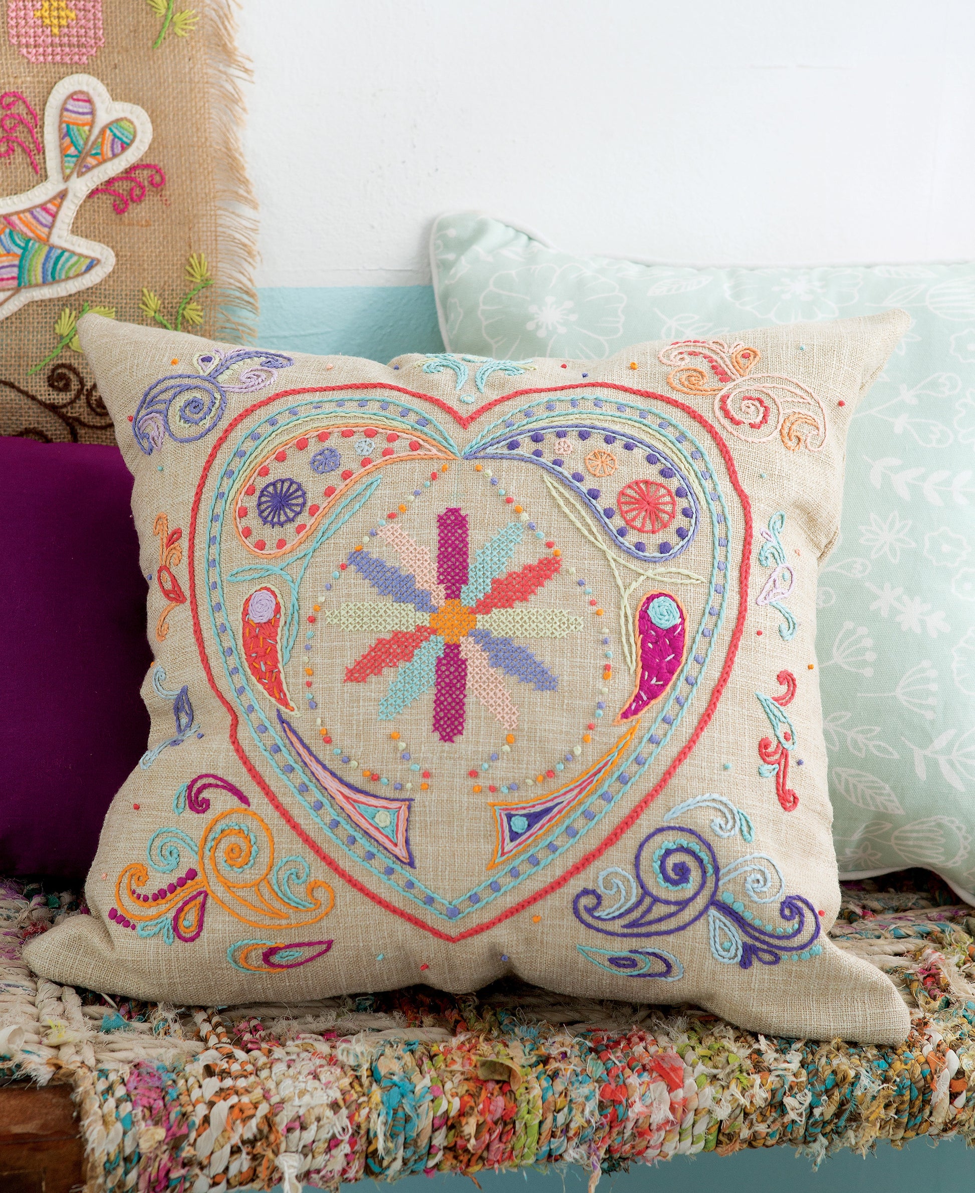 The “Free Spirit Stitching” decorative pillow by Leisure Arts features colorful, intricate retro Bohemian embroidery with swirling patterns and a heart shape on beige fabric, and is displayed resting on a patterned bench alongside other pillows.