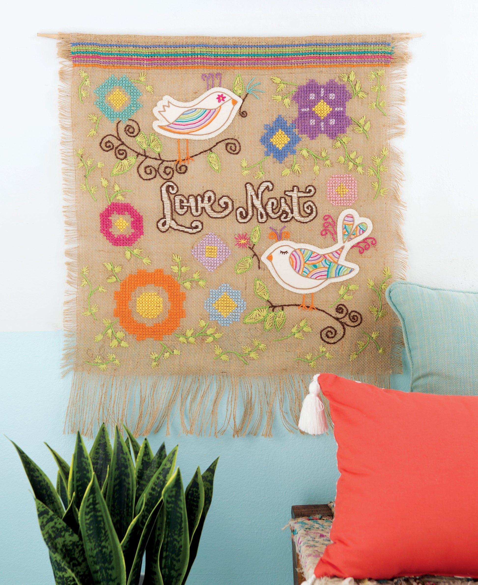 The Free Spirit Stitching wall hanging by Leisure Arts features two colorful birds, vibrant stitched flowers, and the words “Love Nest,” creating a cozy atmosphere. Inspired by retro Bohemian embroidery, it hangs above a green potted plant and an orange pillow against a blue-and-white wall.