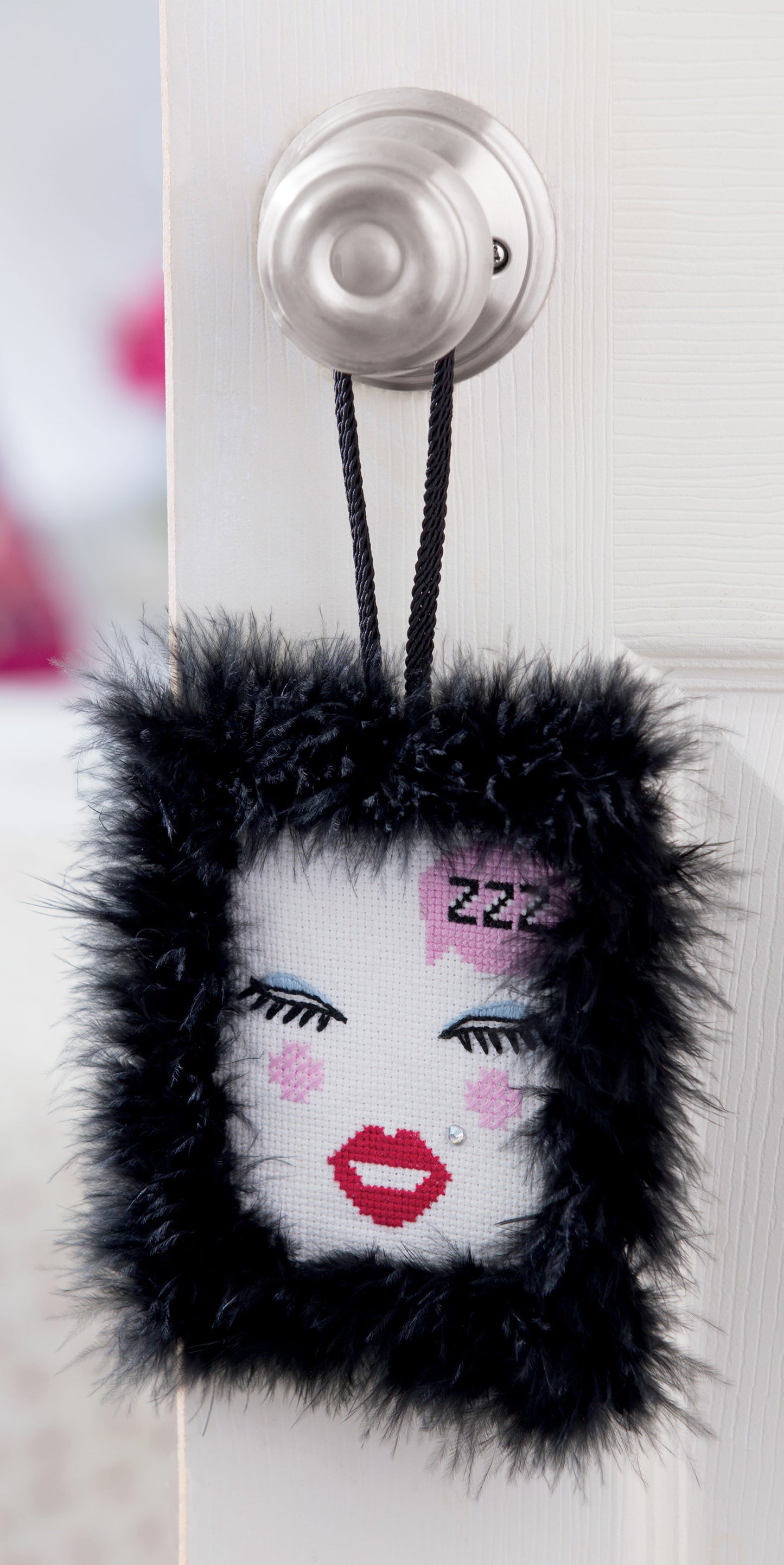 Add whimsical charm to your space with Free Spirit Stitching by Leisure Arts—a square, plush door hanger featuring a face design with red lips, closed eyes, pink blush, and “Zzz” stitched on the forehead. Framed in black faux fur and suspended from a silver doorknob, this accent piece draws inspiration from retro Bohemian embroidery and modern cross-stitch patterns, making it an eye-catching choice for creative needlework projects.