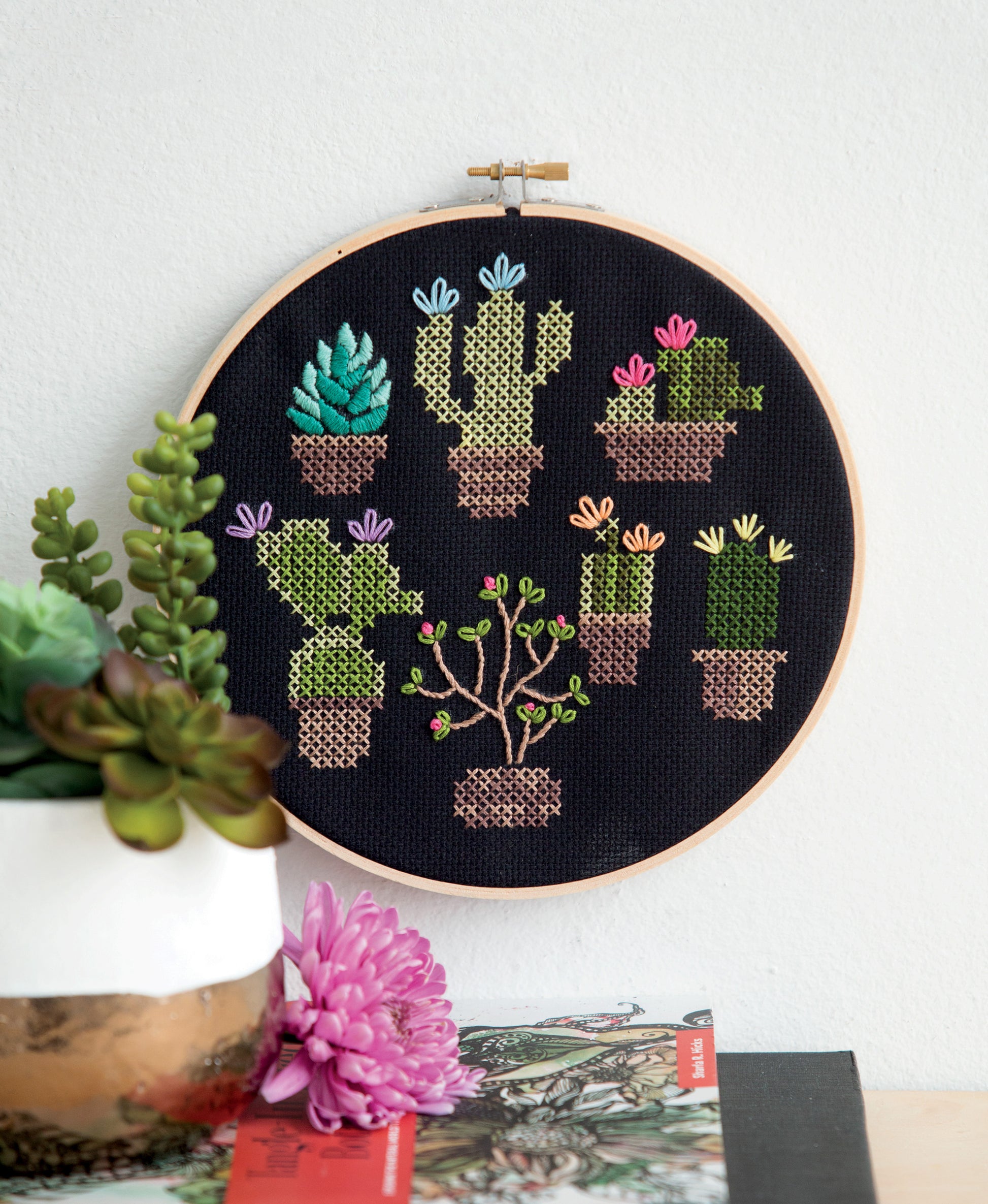 Free Spirit Stitching by Leisure Arts features a vibrant embroidery hoop adorned with cross-stitched potted cacti and succulents on black fabric. Displayed beside a vase of real succulents and a pink flower, this piece showcases retro Bohemian flair—perfect for adding fresh, stylish inspiration to your needlework projects.