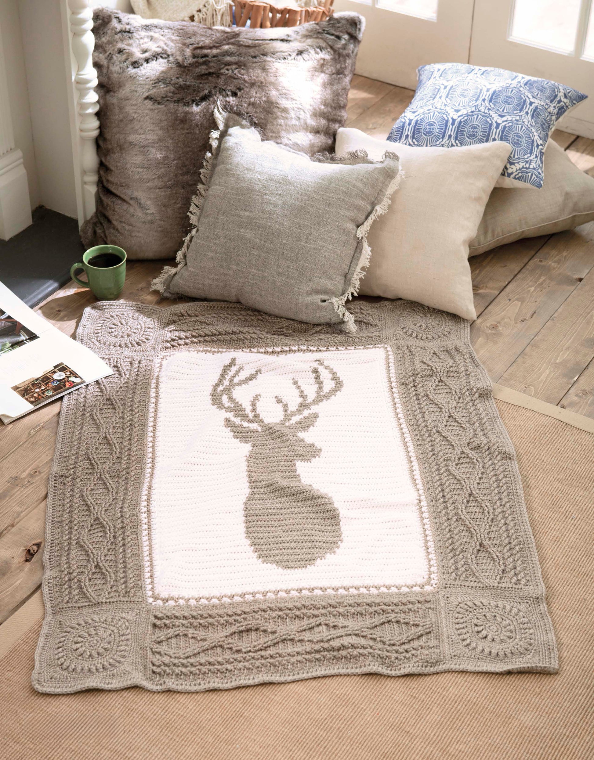 A cozy scene with pillows and a green mug on the floor, featuring the Leisure Arts Wildlife Cable Afghans throw blanket with a deer design, an open book, and soft natural light streaming through nearby doors.