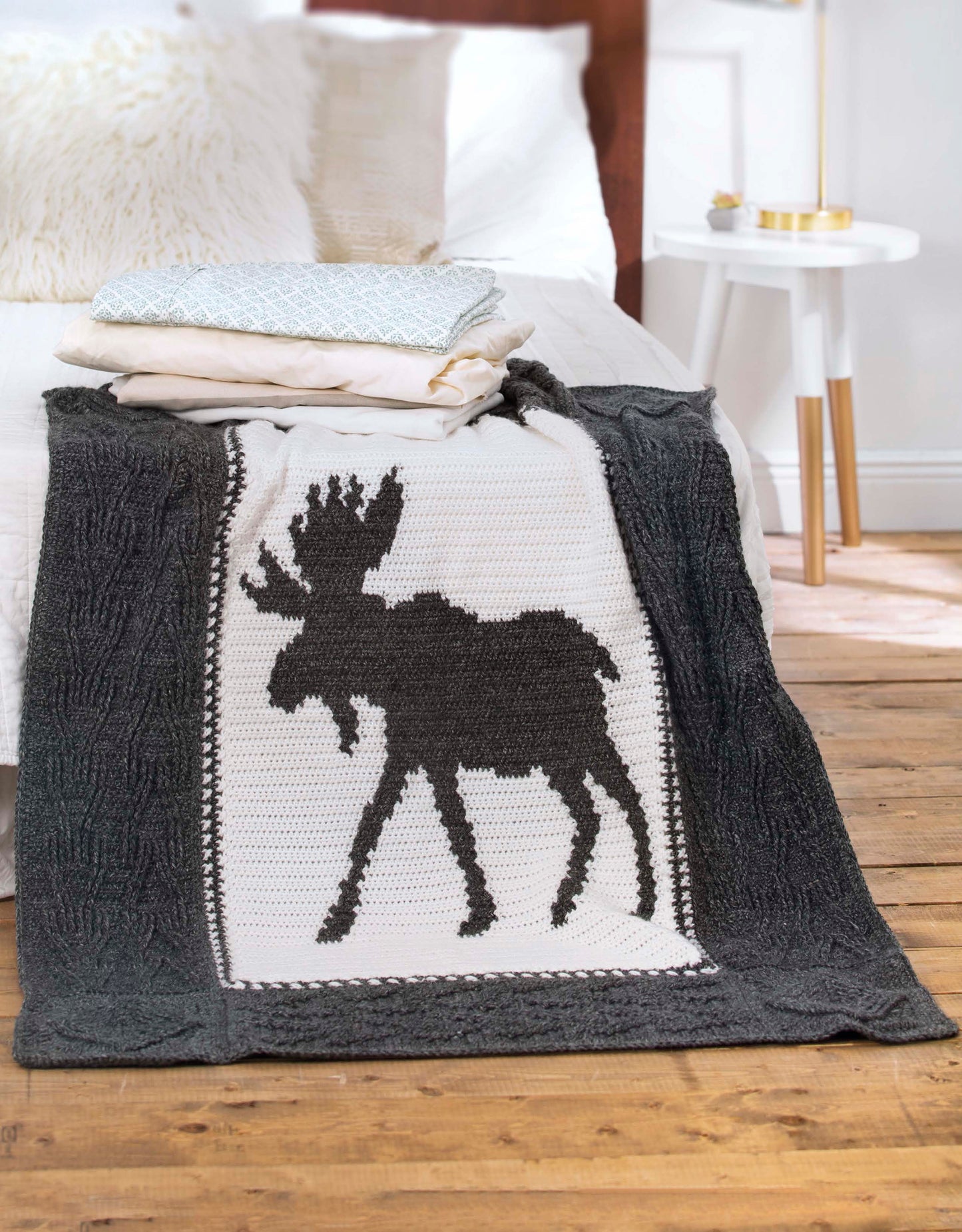 The Wildlife Cable Afghans by Leisure Arts showcase a cozy knit animal throw with a bold moose silhouette, draped over the bed beside folded pillows and throws—ideal for fans of outdoor-inspired crochet décor.