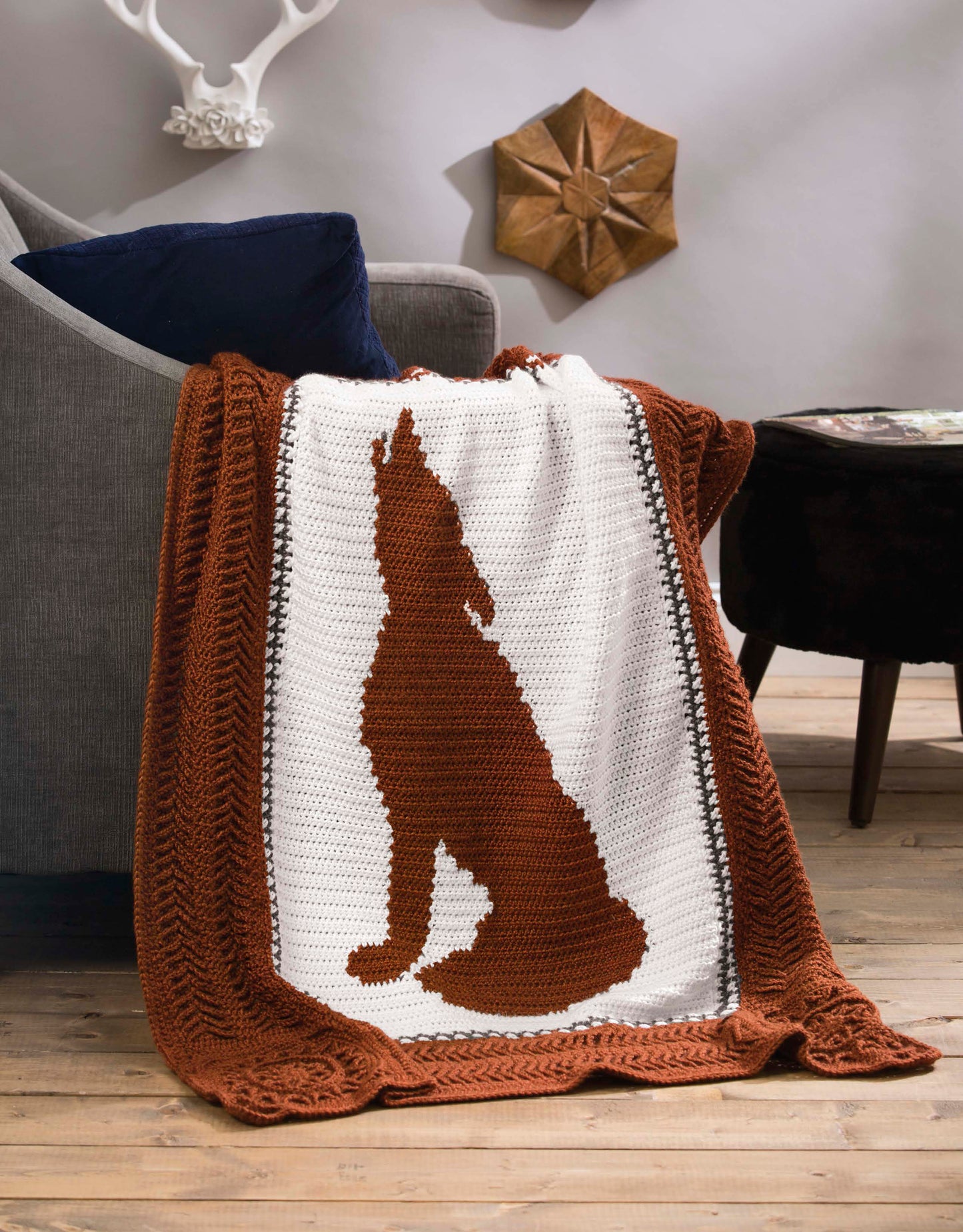 The Leisure Arts Wildlife Cable Afghans crocheted blanket features a large brown howling coyote silhouette on white, adding outdoor-inspired charm to rustic living spaces—perfect for fans of unique animal throws and nature-themed décor.