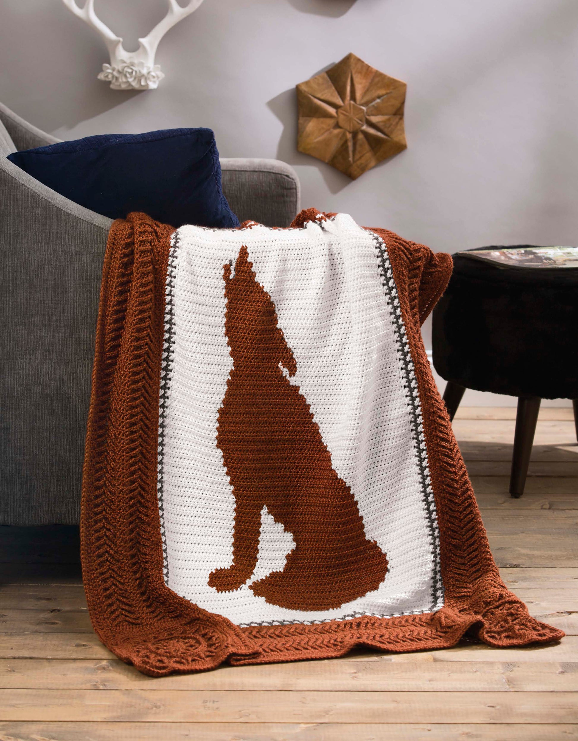 The Leisure Arts Wildlife Cable Afghans crocheted blanket features a large brown howling coyote silhouette on white, adding outdoor-inspired charm to rustic living spaces—perfect for fans of unique animal throws and nature-themed décor.
