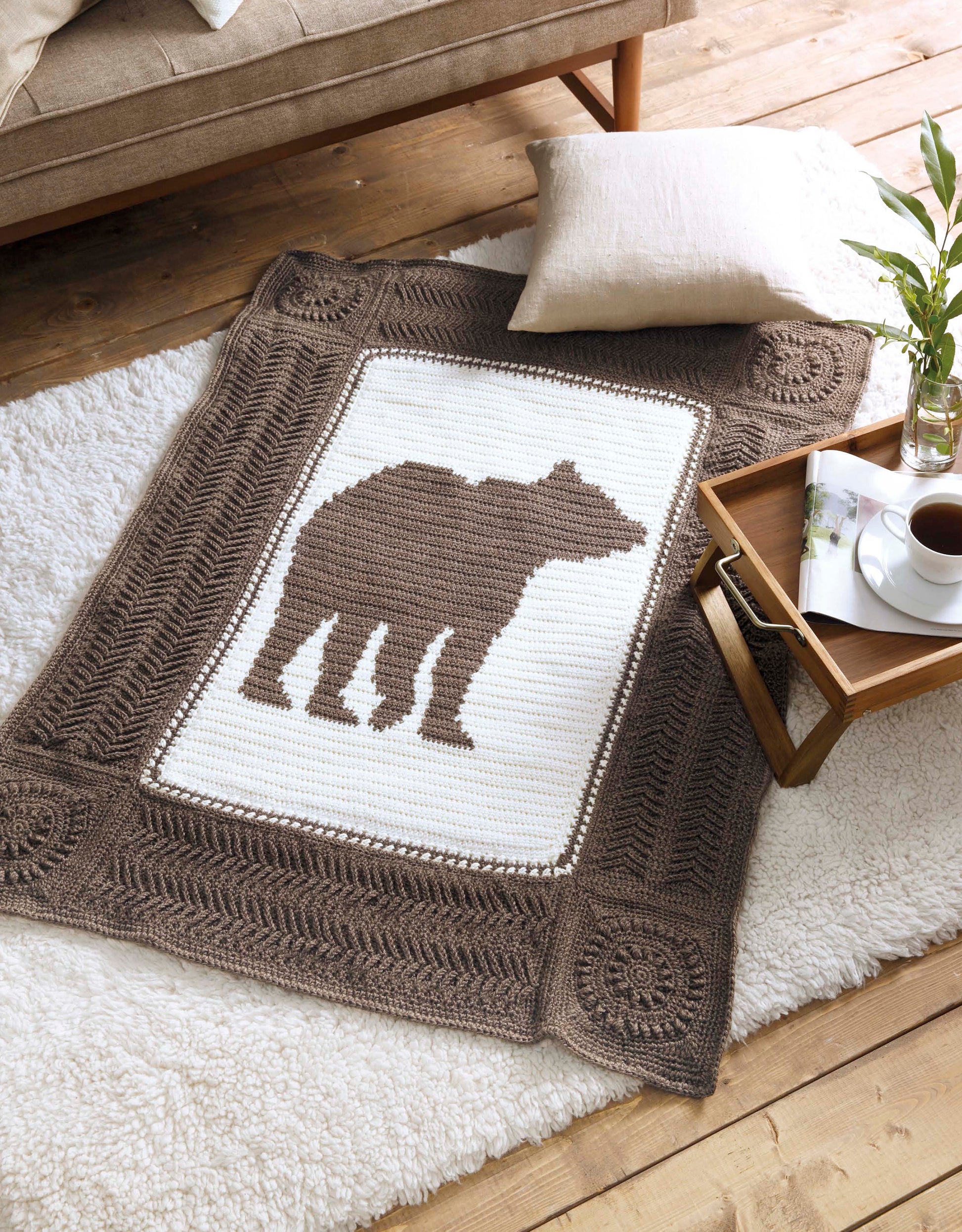 The Wildlife Cable Afghans by Leisure Arts features a crocheted blanket with a brown bear silhouette, shown on a white rug beside coffee and books—ideal for animal-themed decor lovers seeking cozy throw blankets.
