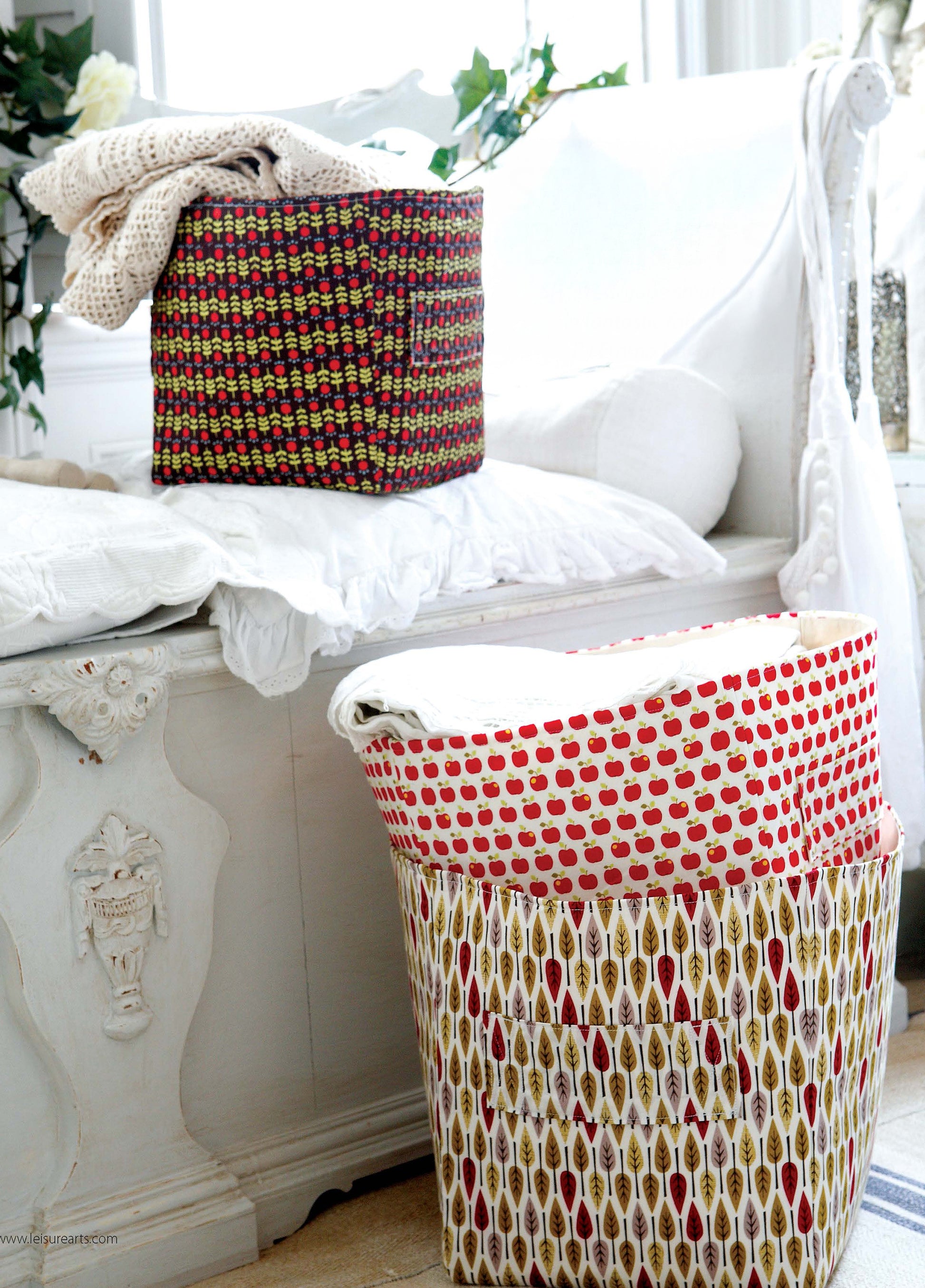 In a bright, cozy room, Sewing Home—11 Useful & Beautiful Decor Projects by Leisure Arts features two patterned laundry baskets beside a white daybed decorated with pillows and a faux fur throw.