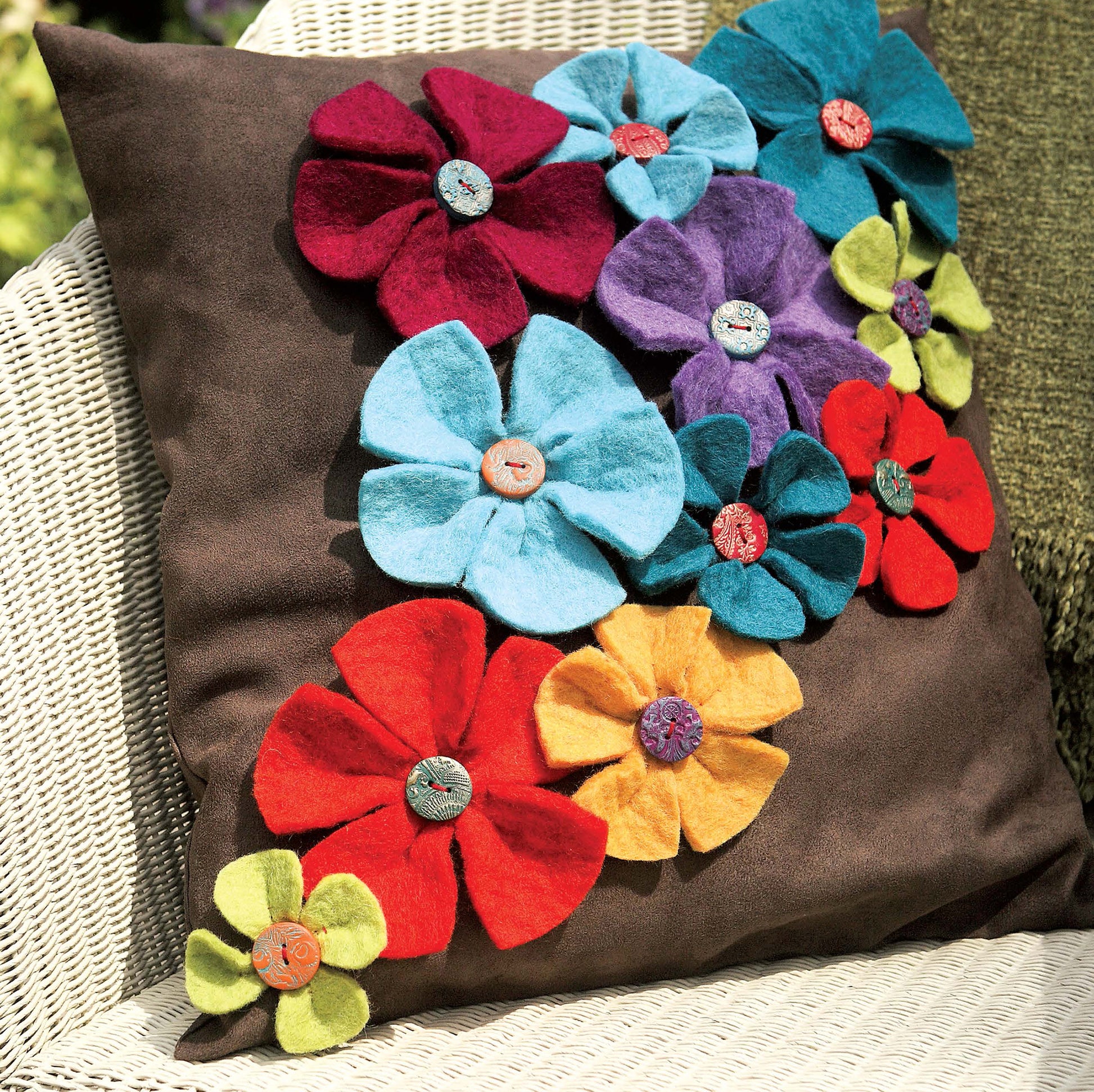 The "Sewing Home—11 Useful & Beautiful Decor Projects" book by Leisure Arts features a brown cushion on a wicker chair, decorated with colorful felt flower corsages in vibrant hues, adding a playful and charming accent to any room.