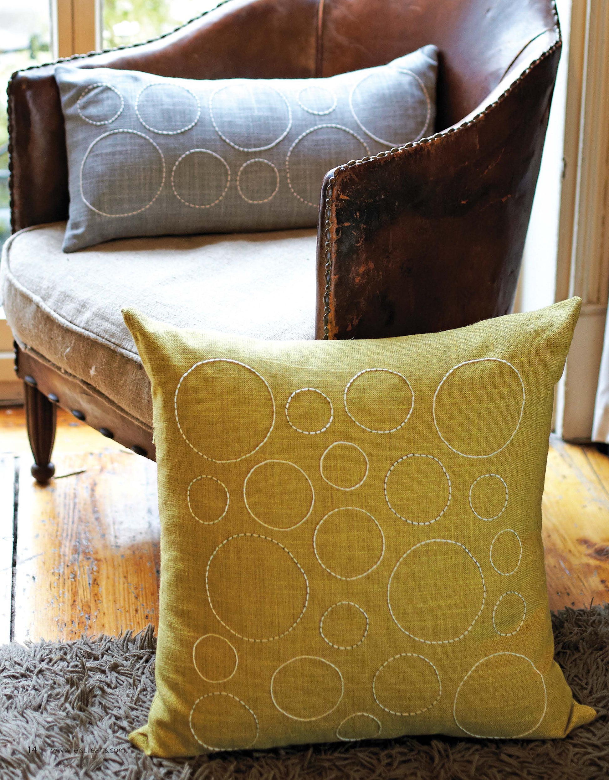 A brown leather chair with a grey pillow sits by a window. A green pillow, both inspired by sashiko Japanese embroidery from "Sewing Home—11 Useful & Beautiful Decor Projects" by Leisure Arts, rests on a rug; both feature circular patterns.