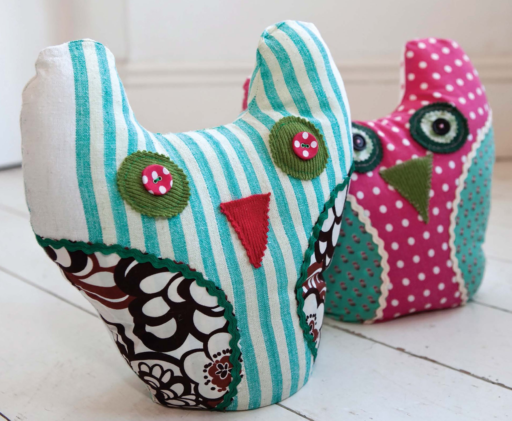 Two handmade fabric owl plushies from Sewing Home—11 Useful & Beautiful Decor Projects by Leisure Arts feature colorful sewn details, button eyes, and sashiko-inspired accents. One has teal stripes and florals; the other is pink with polka dots.