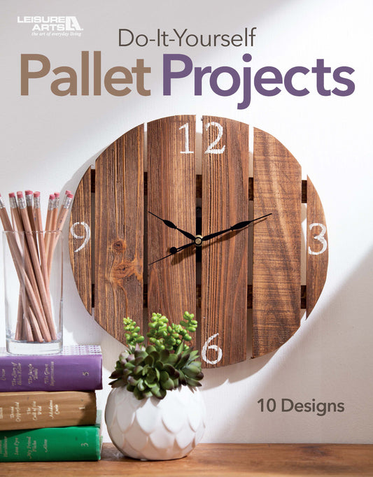 A wooden pallet clock with white numbers hangs above books, pencils, and a potted succulent. The text reads, Do-It-Yourself Pallet Projects by Leisure Arts: 10 designs with wood shipping pallets.
