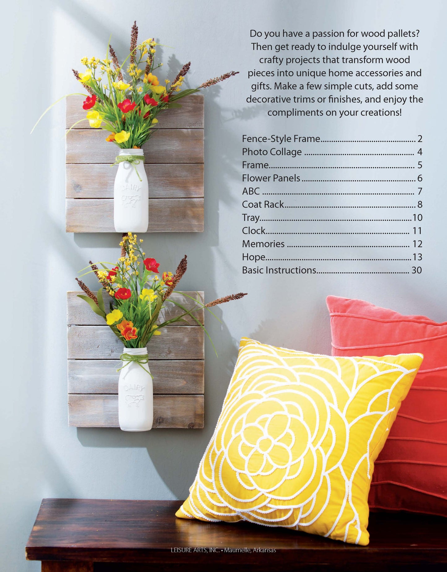 Two wall-mounted wood pallets display white vases of colorful wildflowers, placed beside a yellow pillow with a white floral design and the "Do-It-Yourself Pallet Projects" book by Leisure Arts for creative inspiration.
