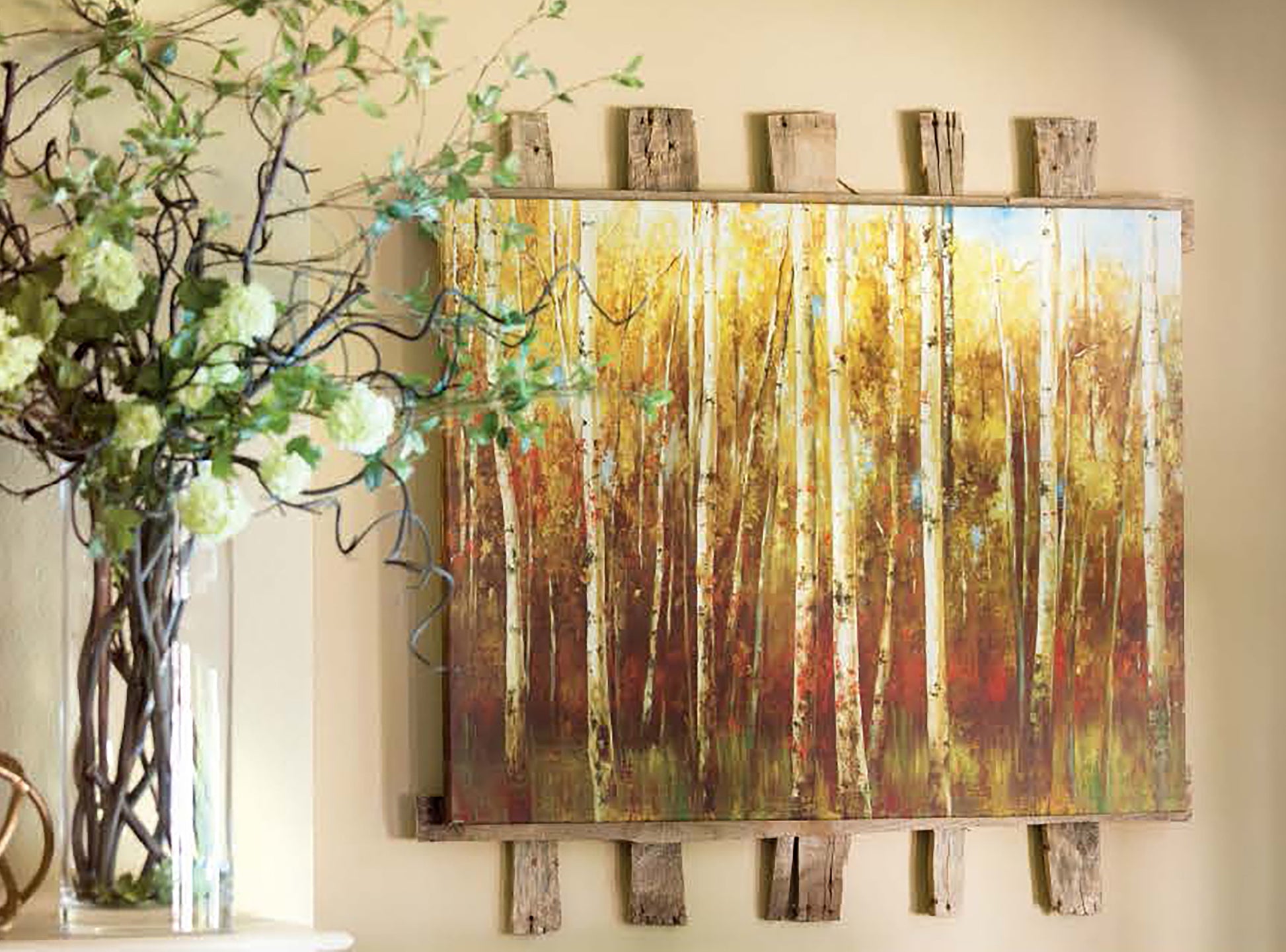 A framed painting of tall trees with yellow and green foliage hangs on a beige wall. To the left, a vase with green and white flowers rests on a white mantel crafted from upcycled pallets using Leisure Arts' Do-It-Yourself Pallet Projects.