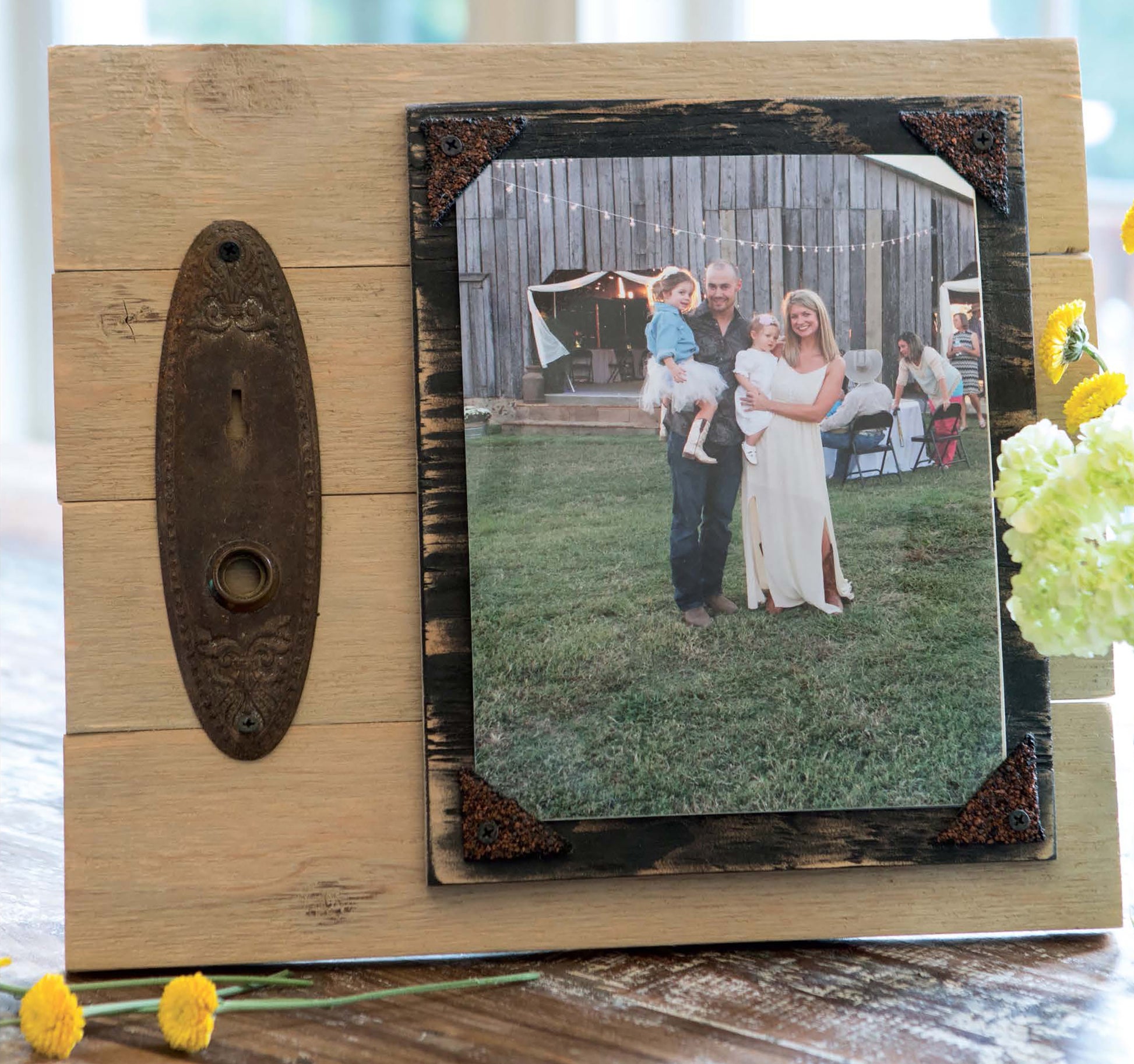 A rustic wooden frame with metal accents, inspired by Leisure Arts’ Do-It-Yourself Pallet Projects, displays a family photo on grass in front of a barn, with yellow flowers in the foreground.