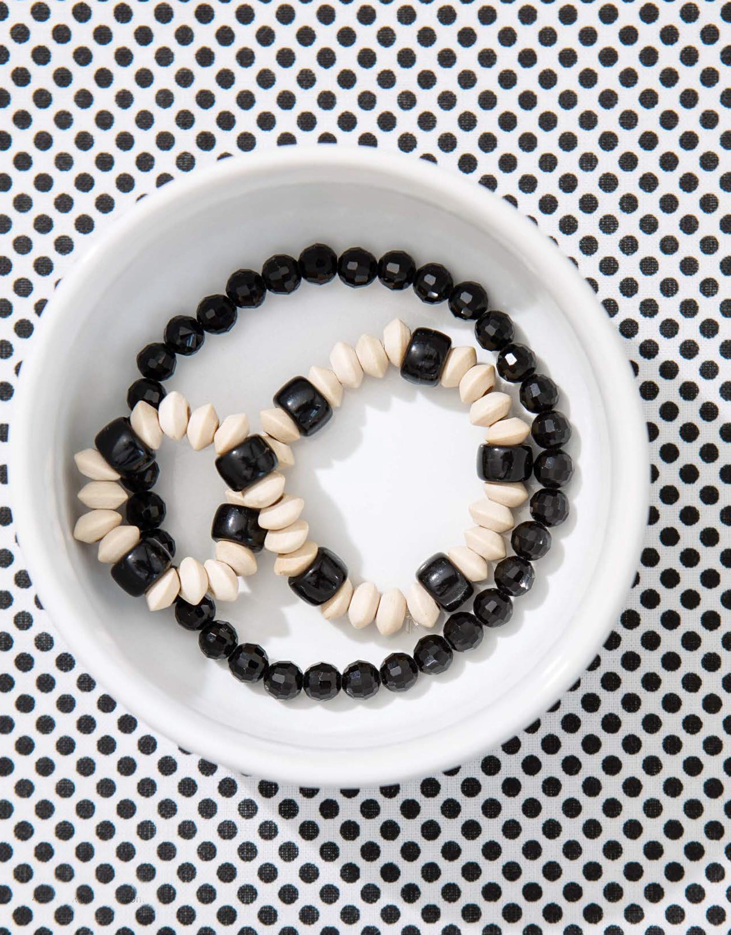 A white bowl holds two beaded bracelets—one black and white, one all black—on a polka dot surface. Quick Fun Jewelry Digital Download by Leisure Arts is perfect for beginners or anyone who loves easy craft projects.