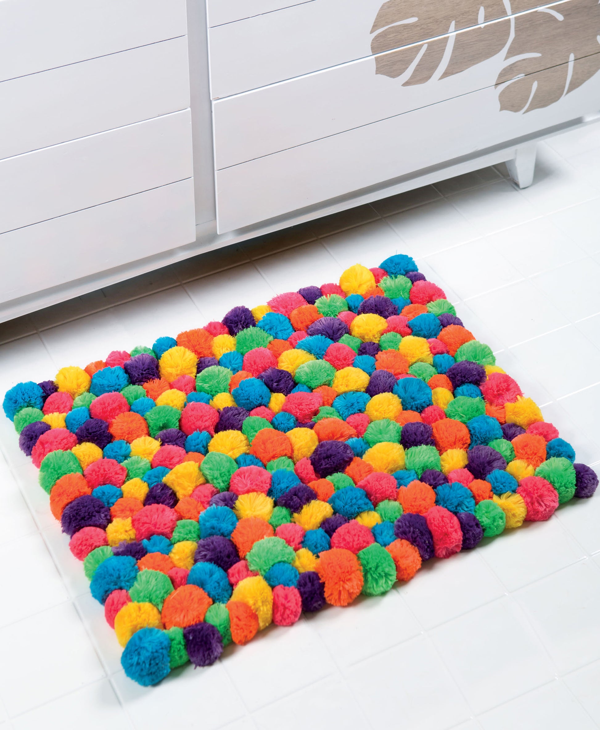 Add a touch of color to your home with this vibrant mat crafted from round, fluffy pom-poms in various bright hues, displayed here on a white tiled floor in front of a white dresser adorned with leaf designs. Pom-Pom Crafts by Leisure Arts is an easy DIY home decor idea—perfect for beginners or anyone eager to explore creative pom pom projects!.