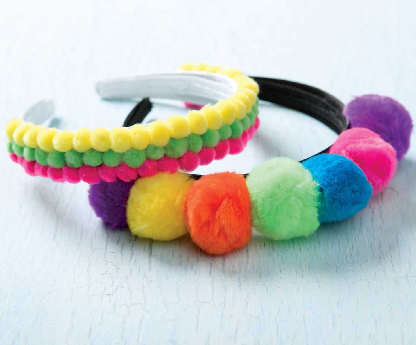 Two colorful headbands from Leisure Arts’ Pom-Pom Crafts collection are displayed on a light blue wooden surface. Ideal for anyone interested in easy DIY home decor or pom-pom crafts, one headband is adorned with small yellow, green, and pink pom-poms, while the other showcases larger pom-poms in cheerful rainbow hues.