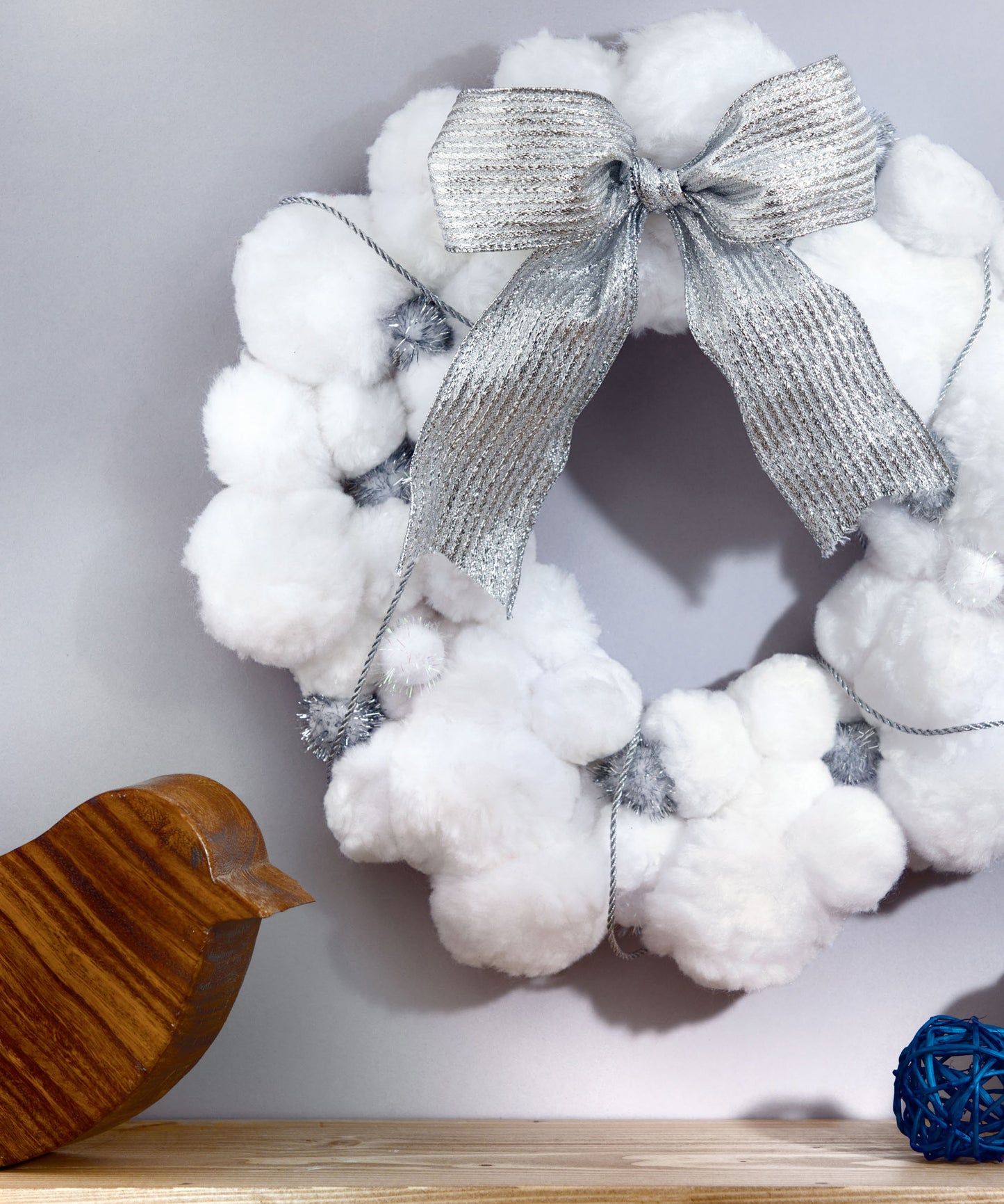 A white pom-pom wreath crafted from fluffy material with a silver ribbon bow by Leisure Arts hangs on the wall. Perfect for easy DIY home decor, this Pom-Pom Crafts project features a wooden bird and a blue decorative ball displayed on the wooden surface below.