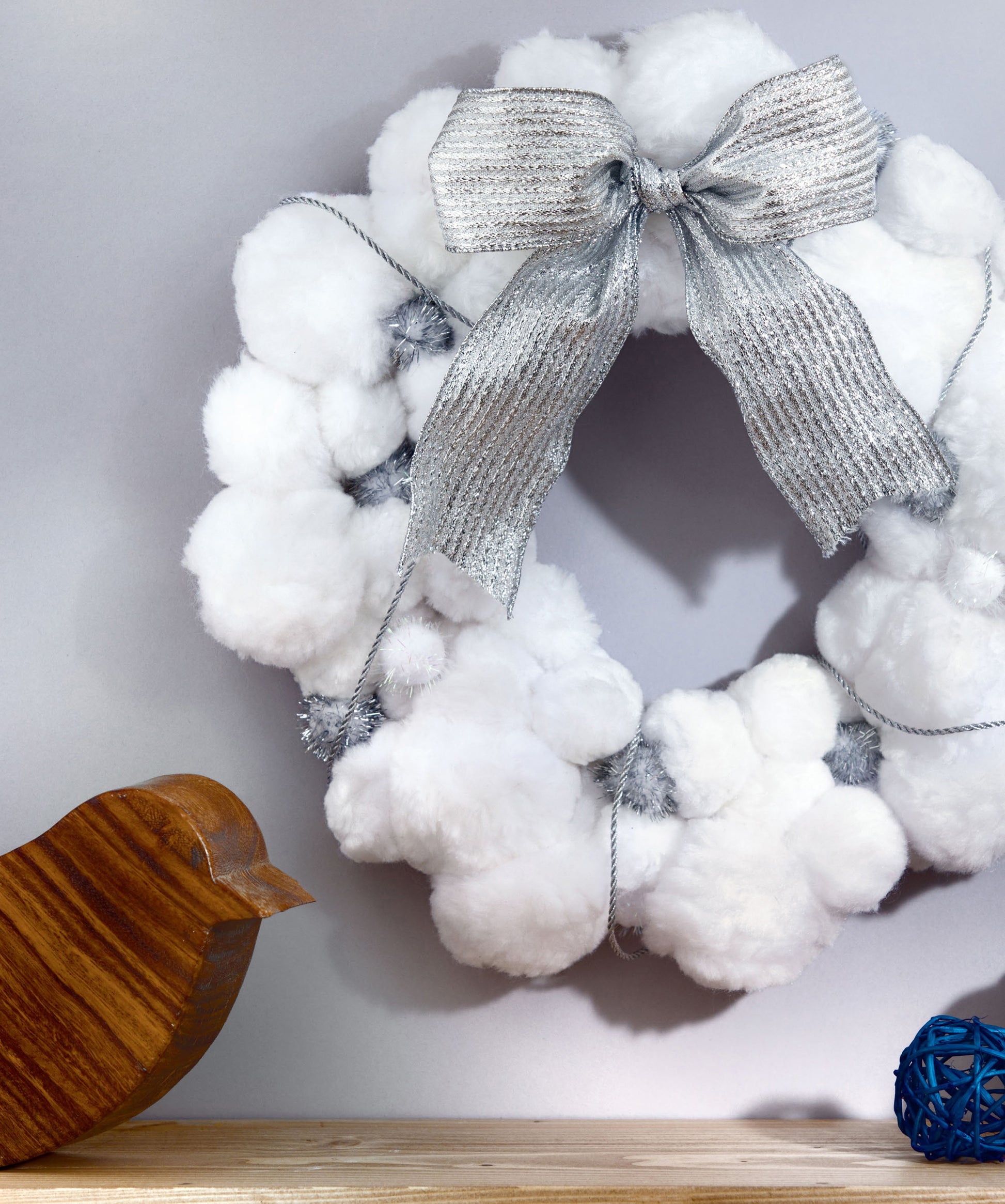 A white pom-pom wreath crafted from fluffy material with a silver ribbon bow by Leisure Arts hangs on the wall. Perfect for easy DIY home decor, this Pom-Pom Crafts project features a wooden bird and a blue decorative ball displayed on the wooden surface below.